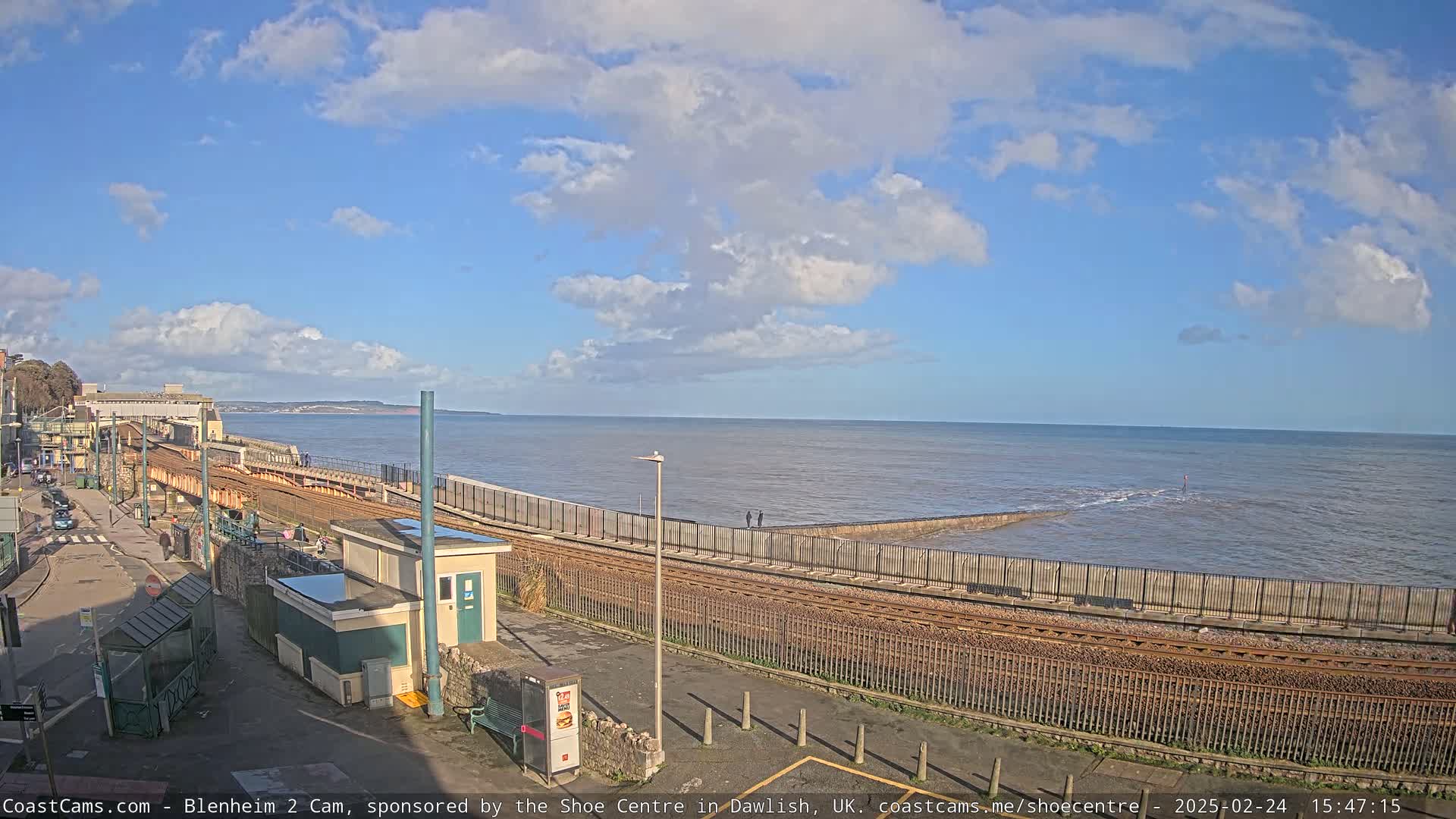 A coastal train track runs alongside a sea wall, with a calm ocean under a partly cloudy sky.