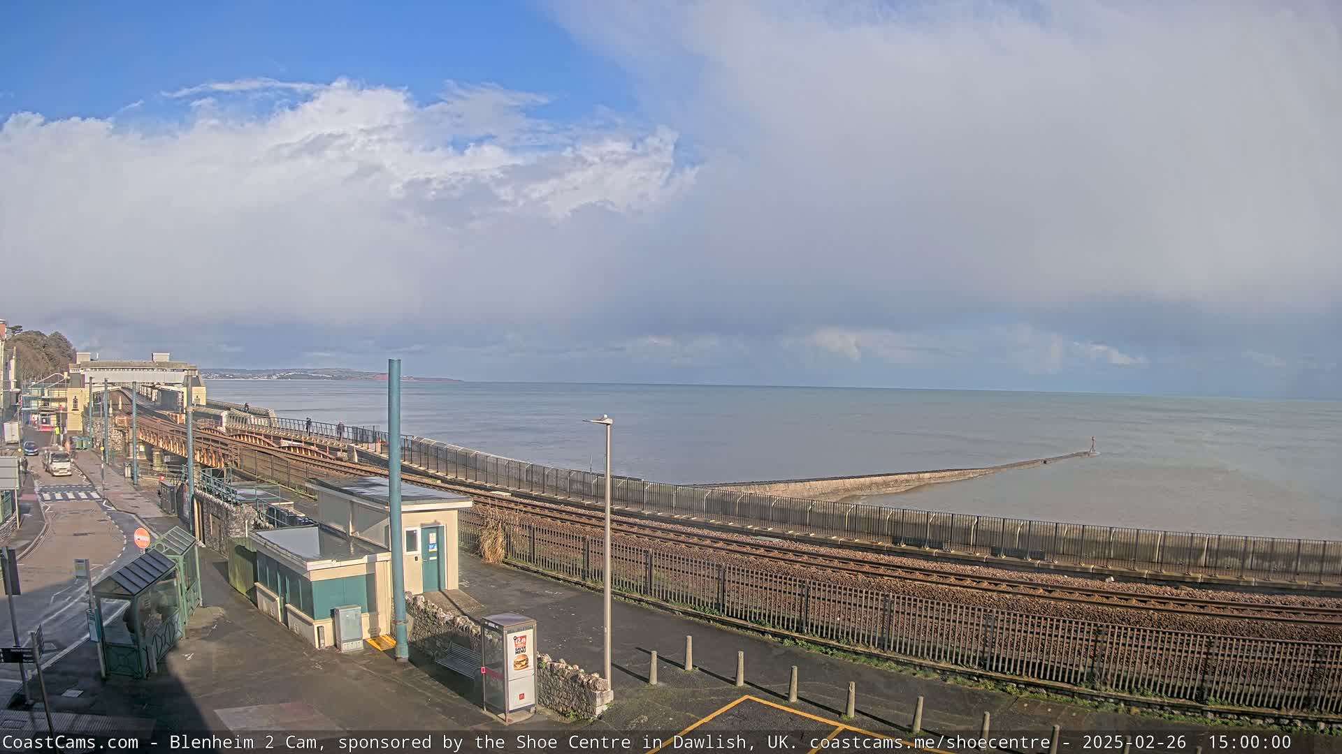 A mostly cloudy day reveals a coastal scene with a railway line running alongside a sea wall extending into a calm sea.