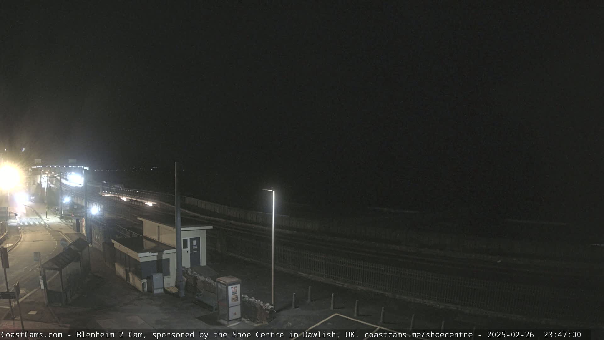 A nighttime view of a coastal area showing a train track, buildings, and a dark, calm sea under a clear, black sky.