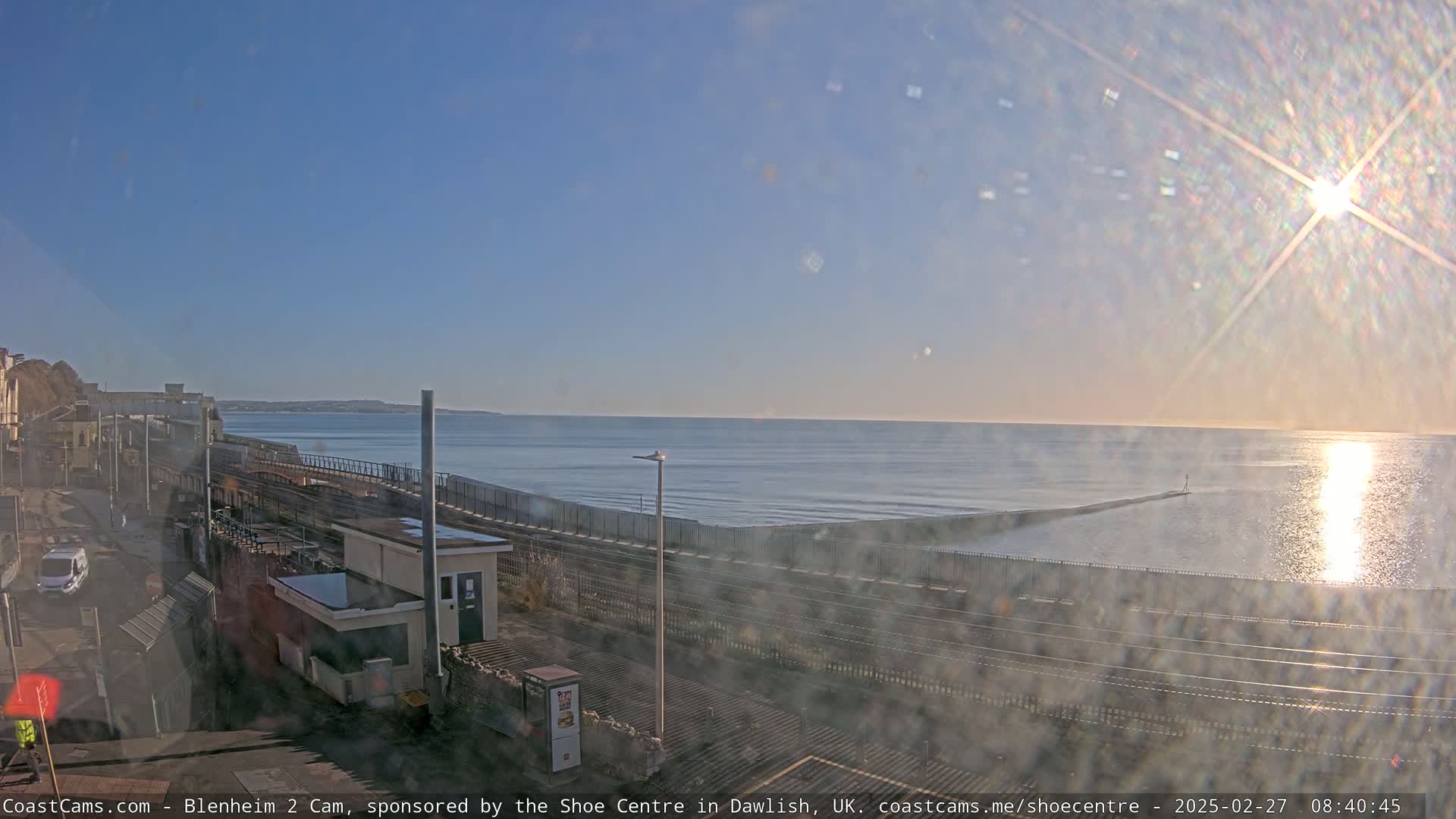A sunny day reveals a coastal scene with train tracks running alongside a sea wall, overlooking a calm ocean and a distant shoreline.