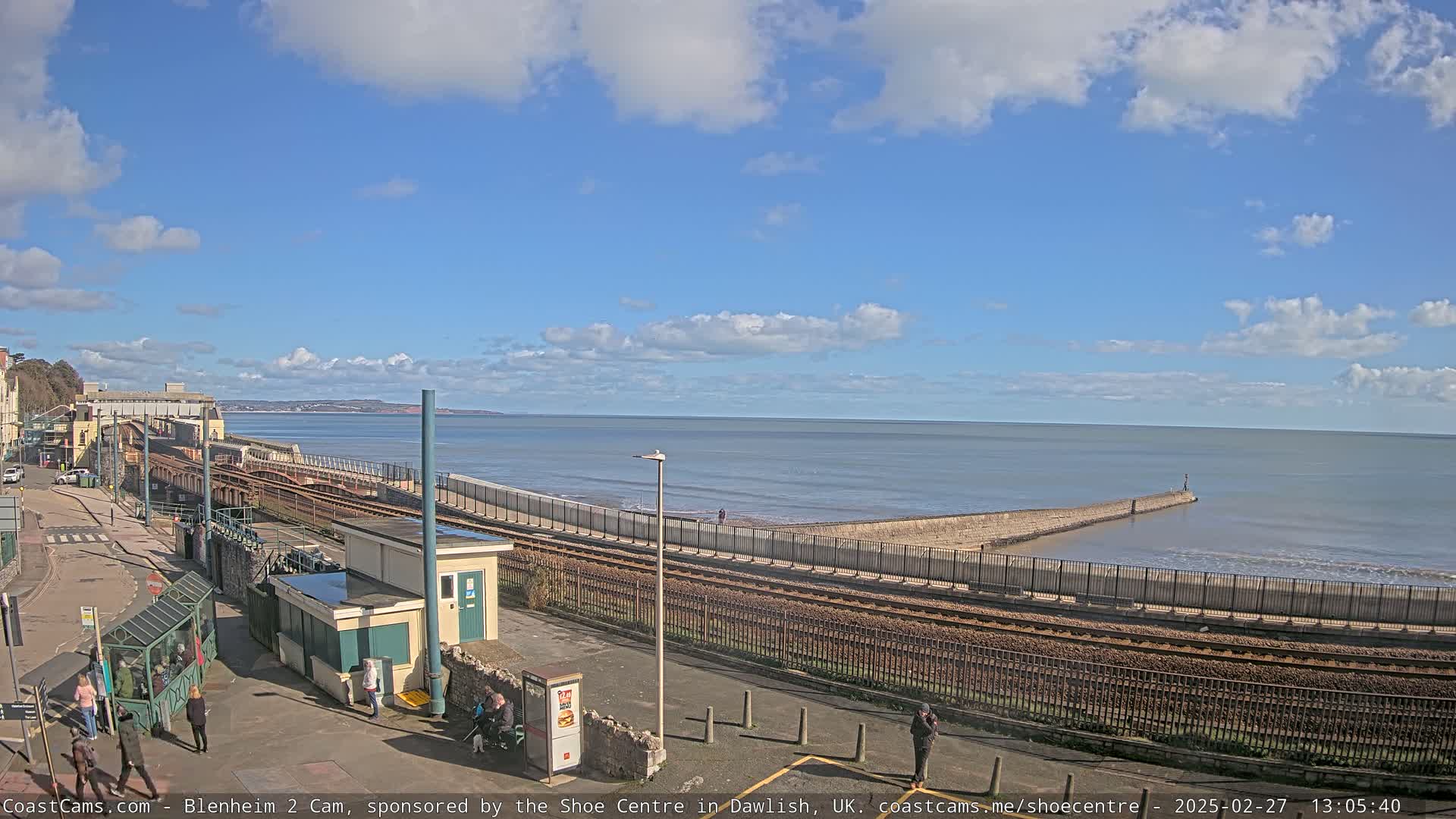 A seaside train track runs along a seawall next to a calm ocean under a partly cloudy sky.