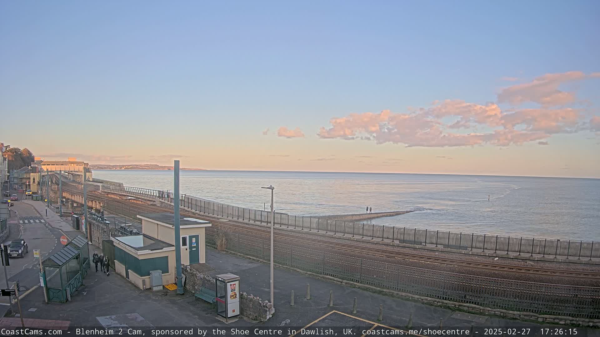 A coastal train track curves alongside a calm sea under a pale pink and blue sky.
