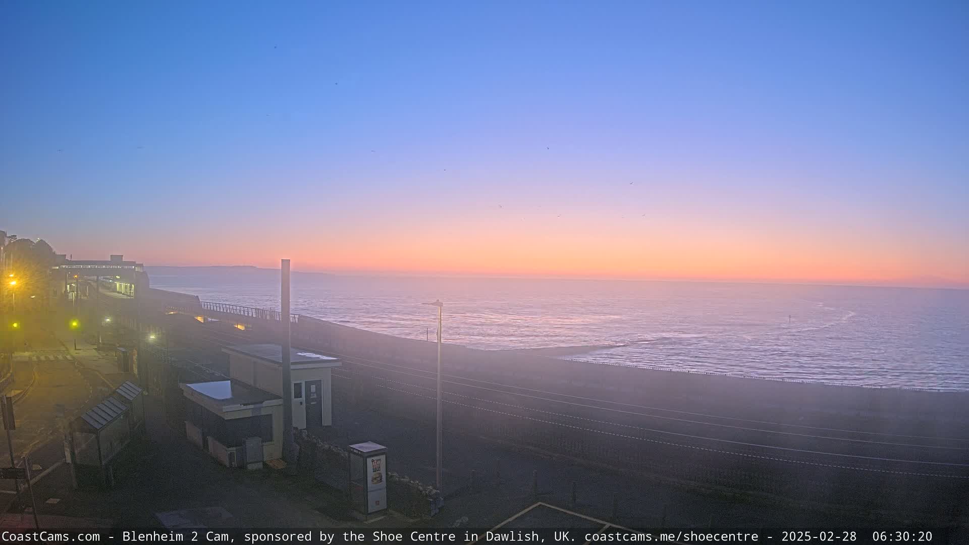 A misty sunrise casts a pinkish-orange glow over a calm ocean, viewed from a coastal area with a building and a rail line in the foreground.