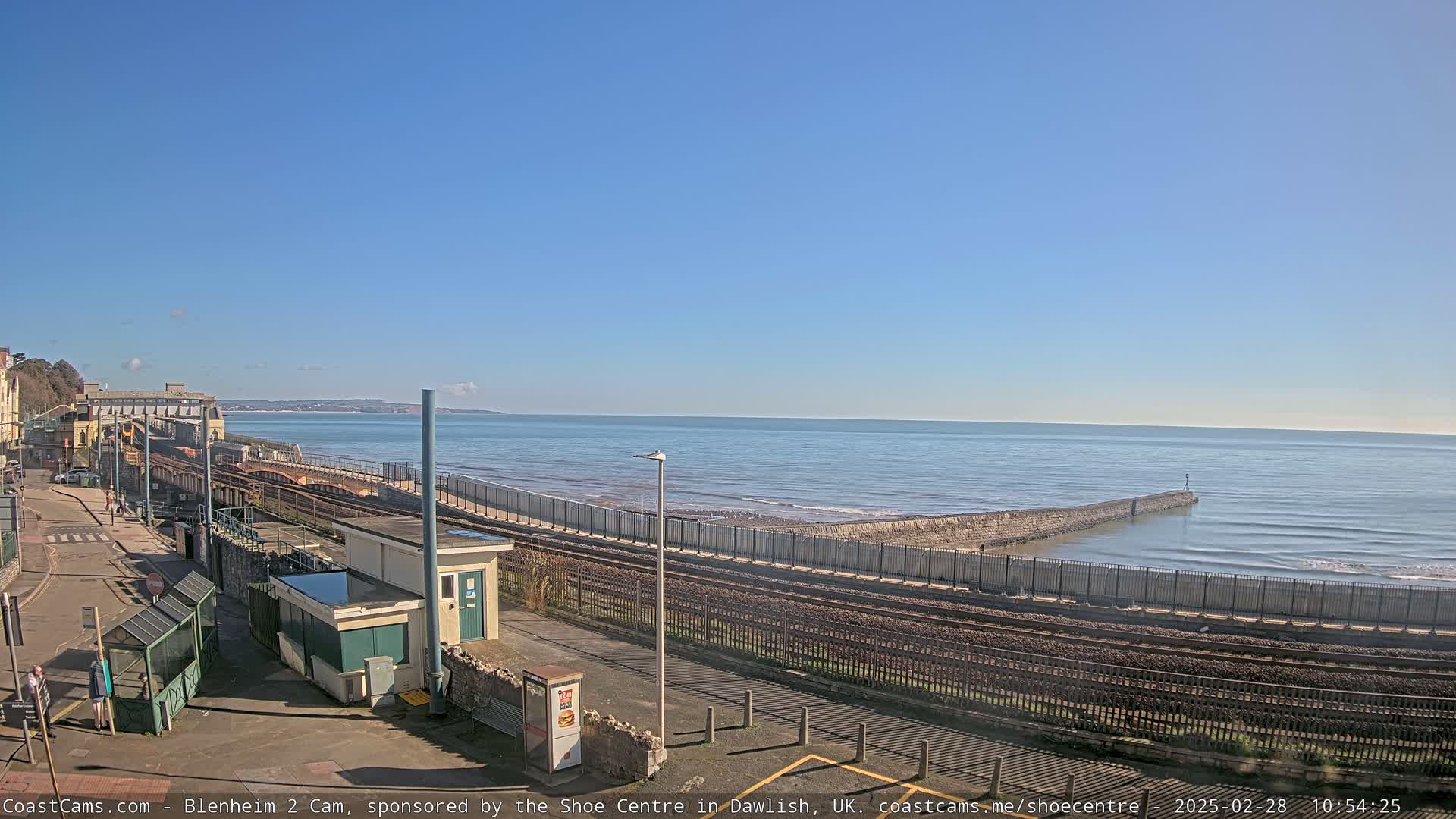 A coastal train station with tracks running alongside a sea wall, overlooking a calm ocean under a clear blue sky.