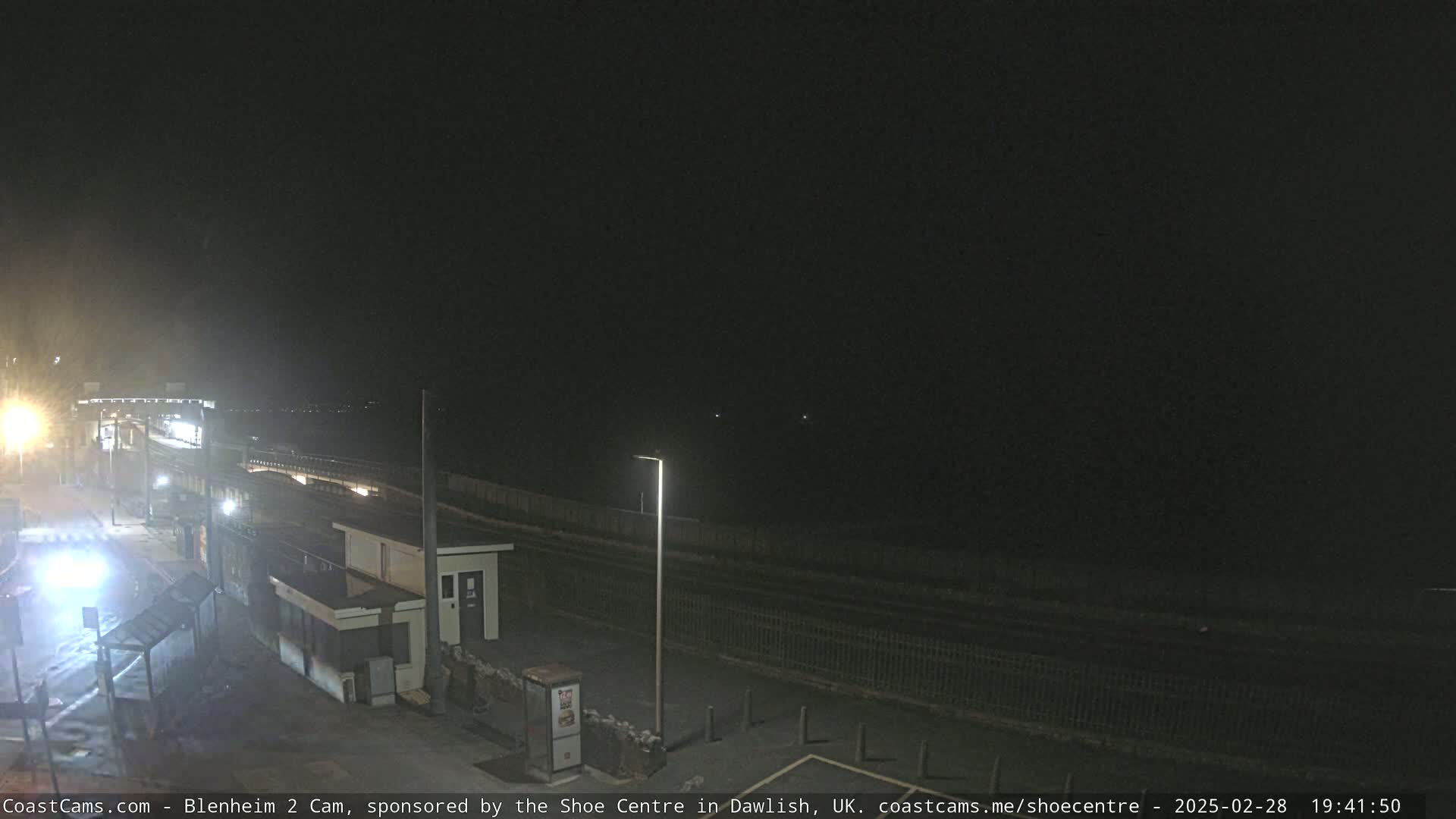 A nighttime view of a mostly empty coastal area with a streetlight illuminating a small building and a section of road next to a fence.