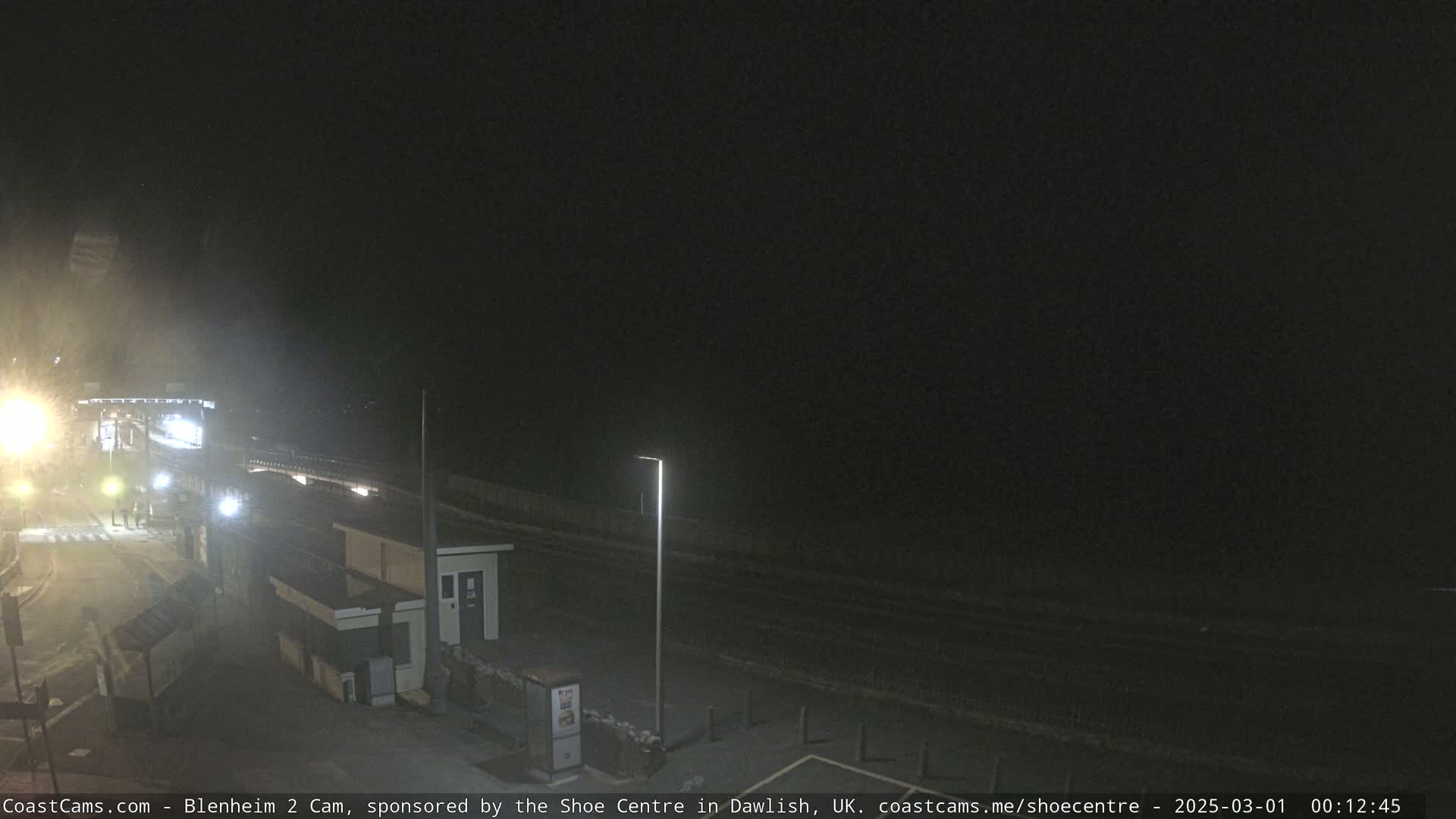 A nighttime, dimly lit coastal scene shows a road, buildings, and a few people near a light post.