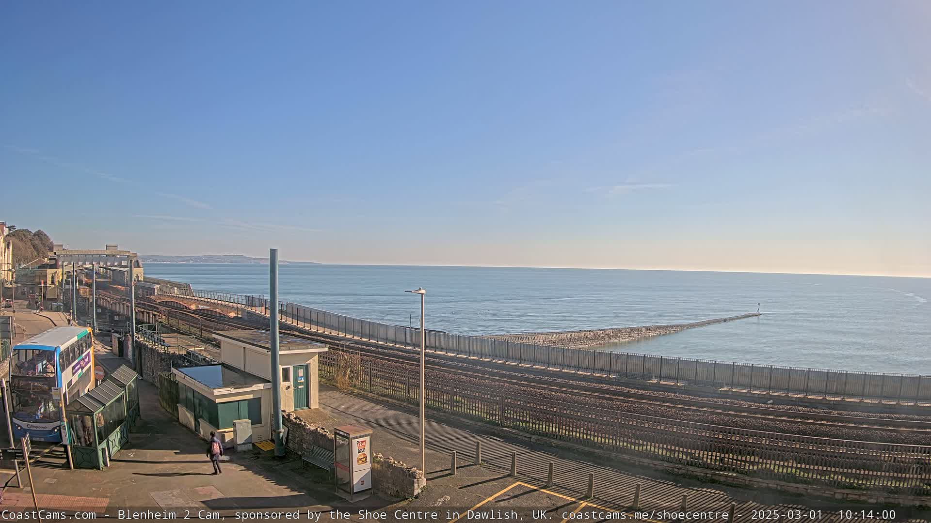 A seaside train track runs alongside a calm ocean under a clear blue sky, with a bus and a few people visible near a station.