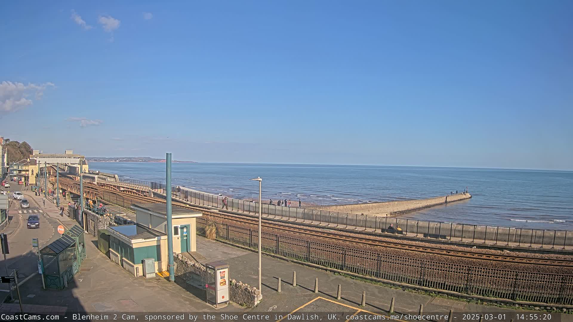 A seaside scene on a sunny day shows a train track curving alongside a paved walkway that extends to a low stone jetty jutting into the ocean.