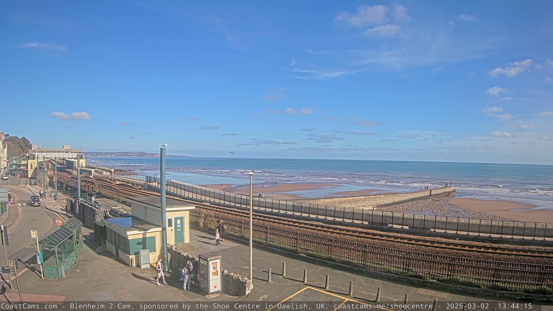A coastal scene on a sunny day shows a train track running alongside a beach with a low wall separating them, several people walking near the track, and a calm ocean beyond.