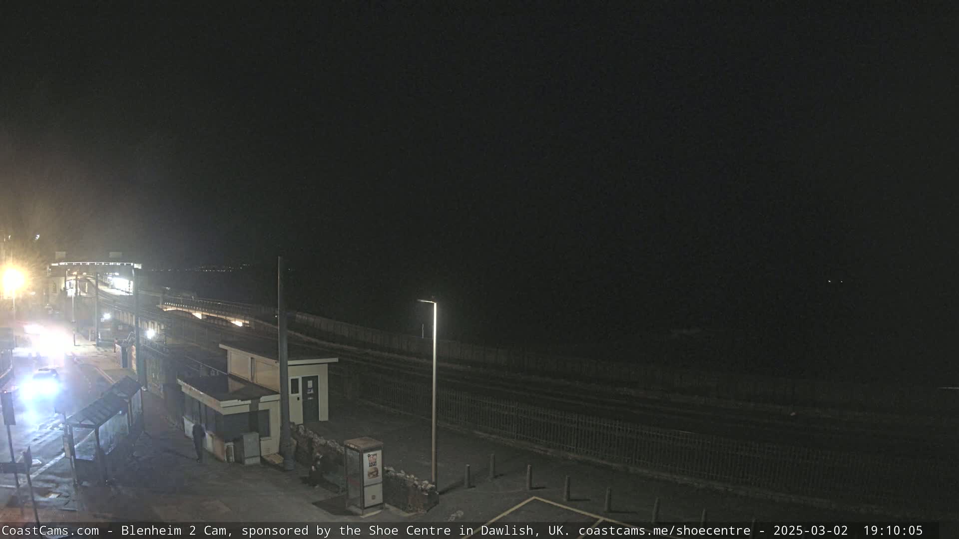 A nighttime view of a seaside area with a paved road, some buildings, and a fence separating the road from the dark ocean.