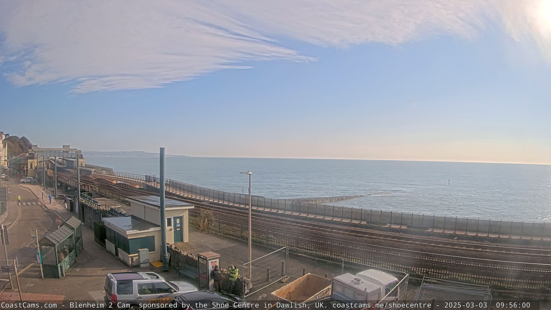 A seaside train track runs along a coastline under a mostly clear, sunny sky with a calm sea.