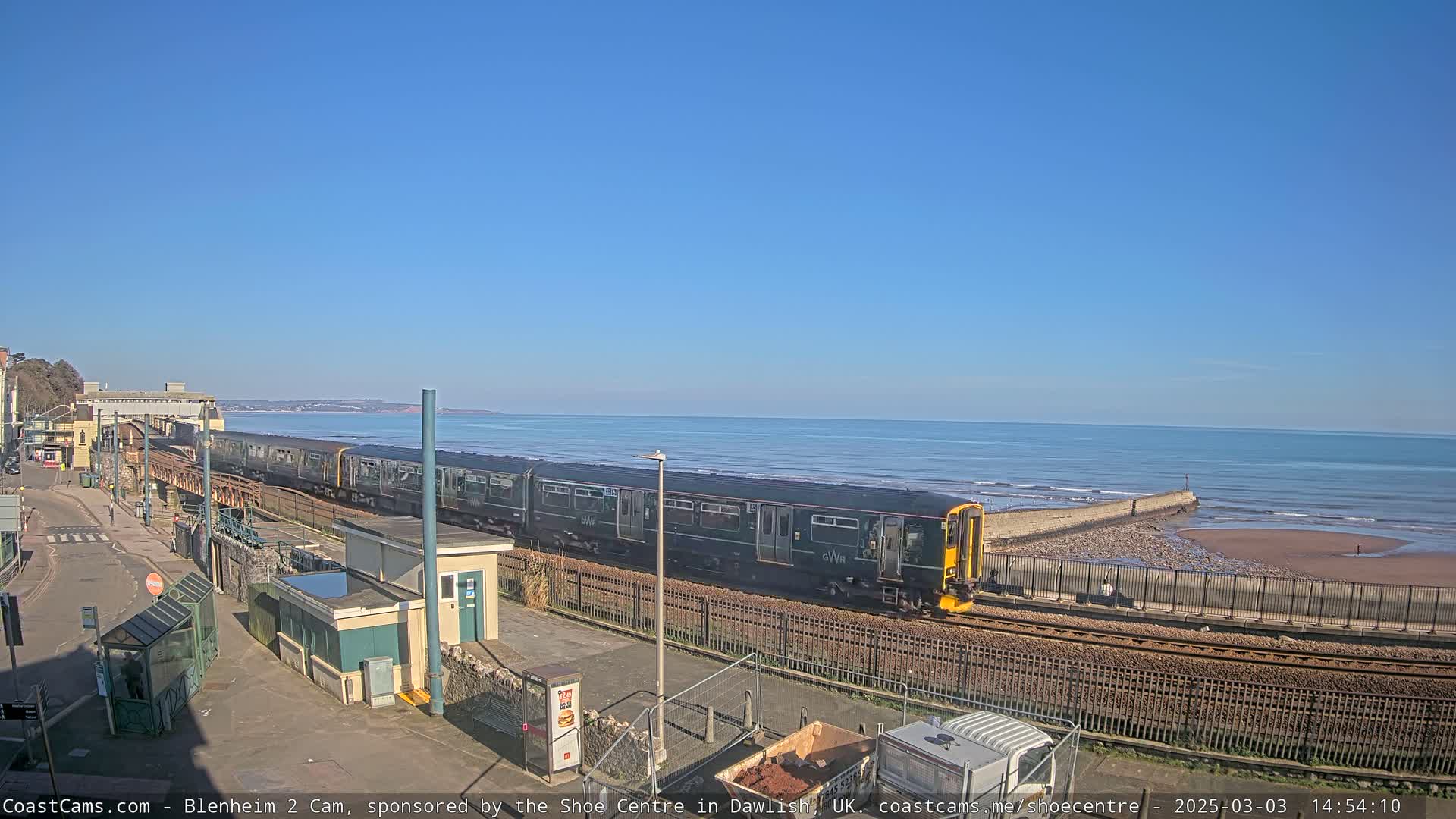 A long dark-green train travels along a coastal railway line on a sunny day, with a calm ocean visible in the background.