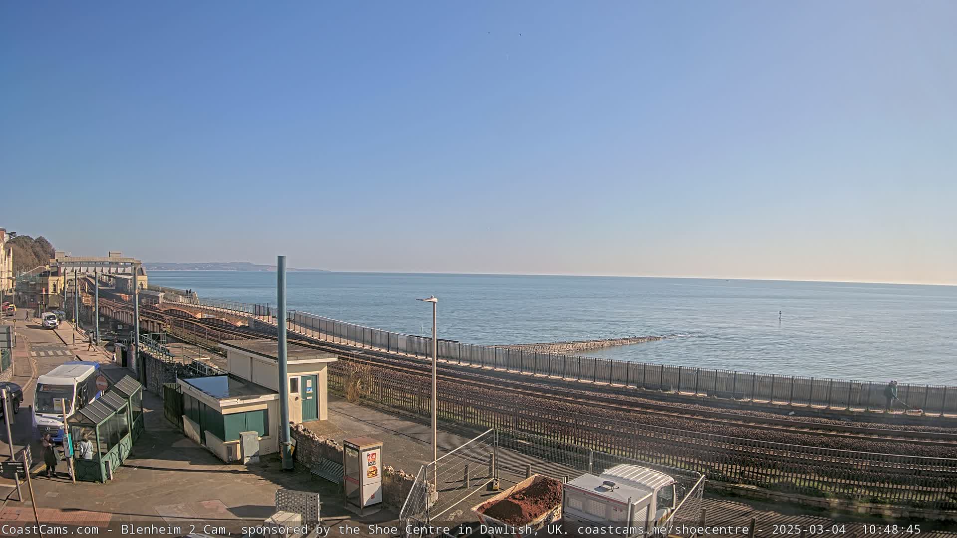 A coastal train station and tracks next to a calm ocean under a clear blue sky.