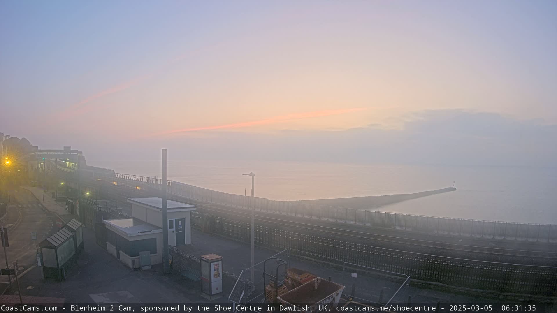 A hazy sunrise over a calm sea, viewed from a coastal railway platform with a small building and a breakwater visible.