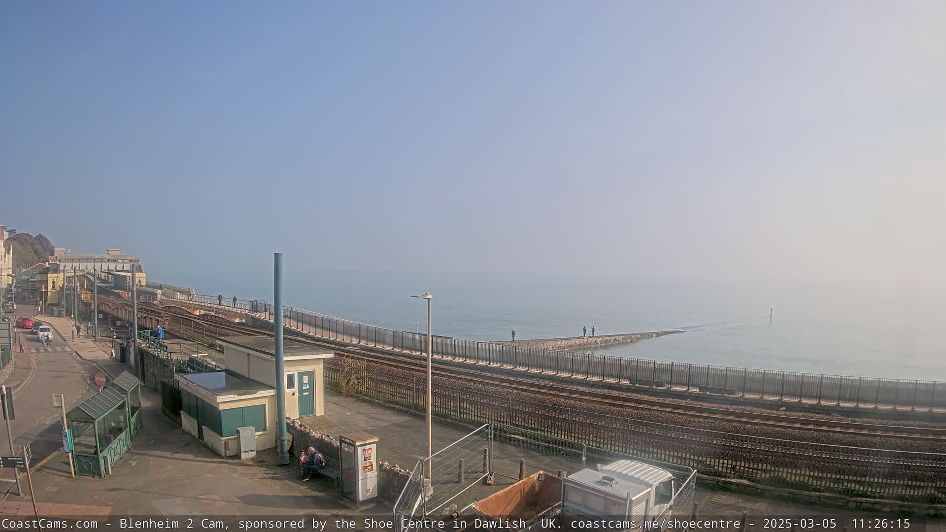 A coastal train station scene on a hazy day, showing train tracks alongside a sea wall with a few people visible, and a small building near the tracks.