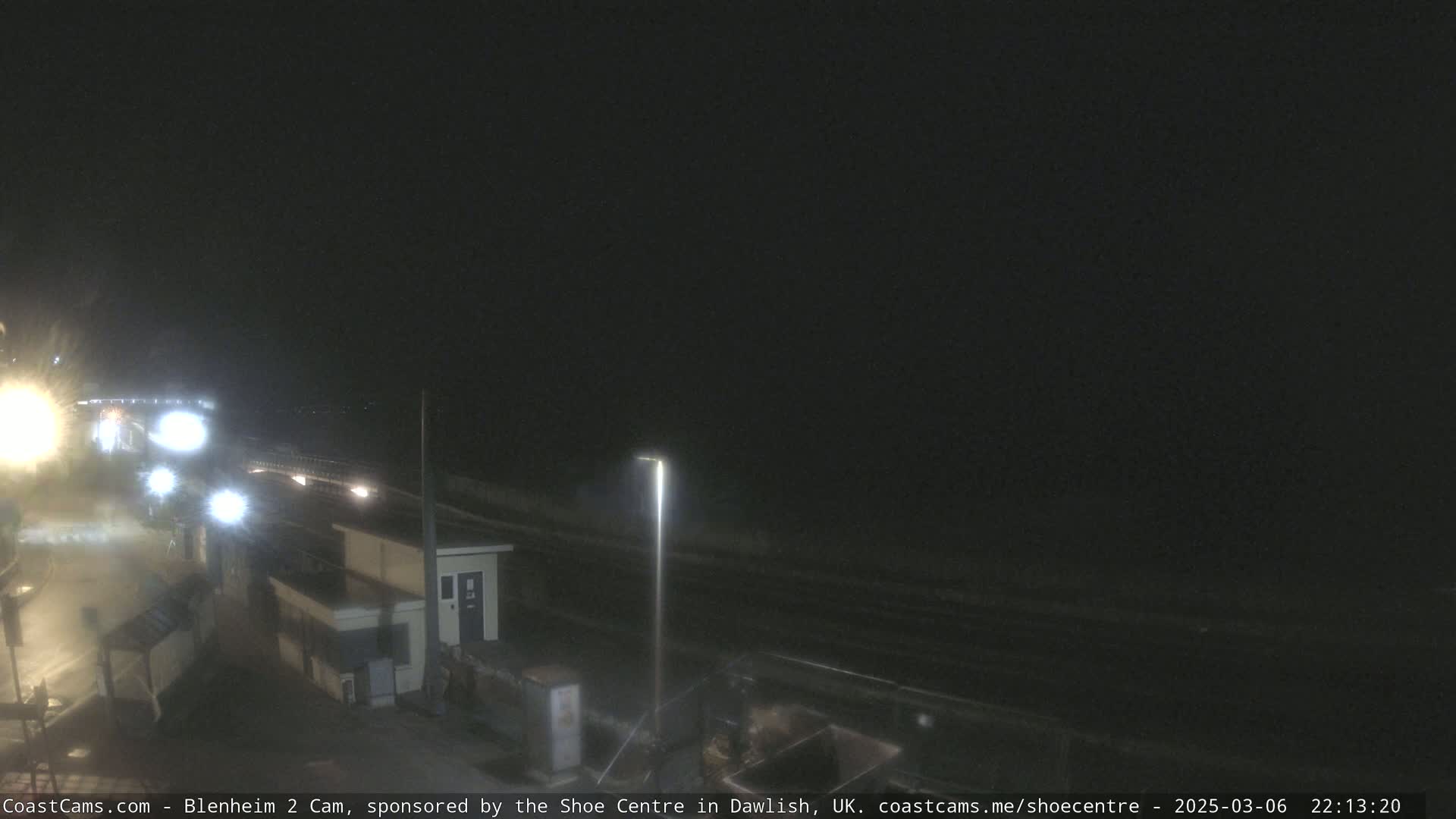 A nighttime coastal scene shows a dark beach and a paved area with streetlights, buildings, and some vehicles.