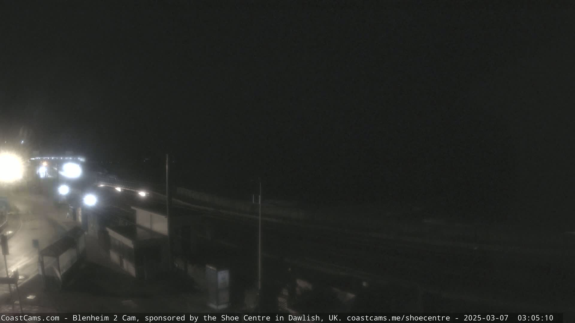 A nighttime, low-light view of a road and shoreline, with some faint vehicle lights visible.