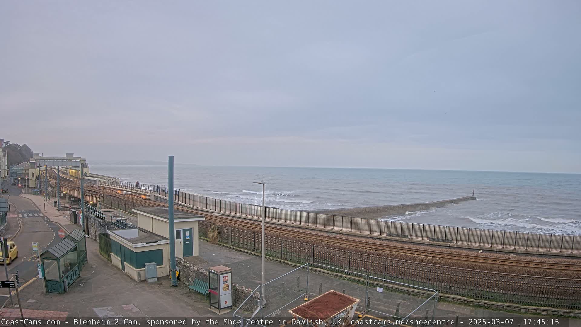 A coastal train track runs alongside a sea wall with moderate waves crashing against it under an overcast sky.