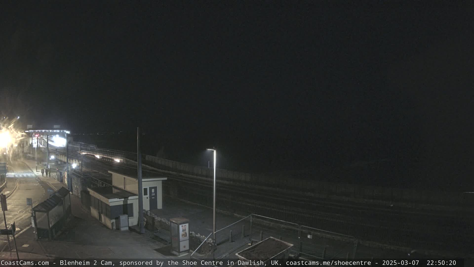 A nighttime view of a mostly empty train platform, with a single light pole illuminating the area and a dark ocean visible in the distance.