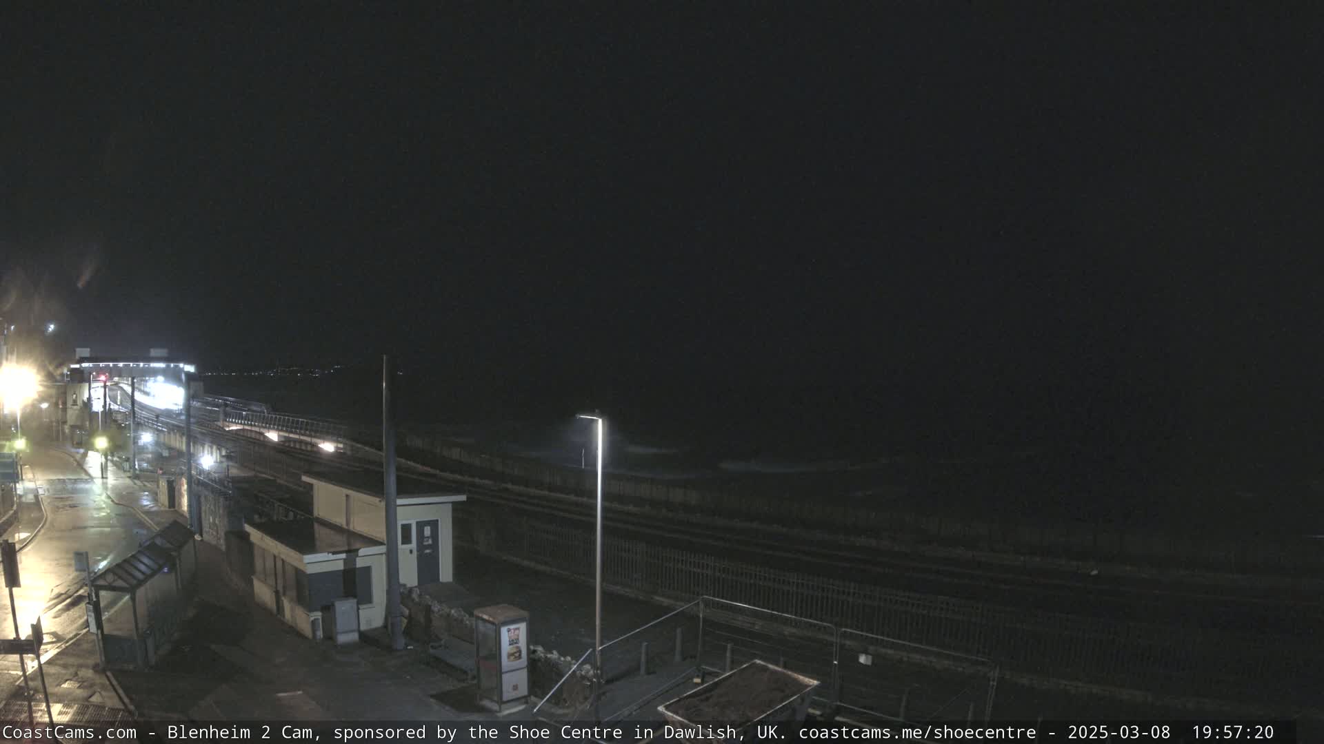 A nighttime view of a wet, dark seaside area with a train track, a small building, and a fence, under a cloudy sky.