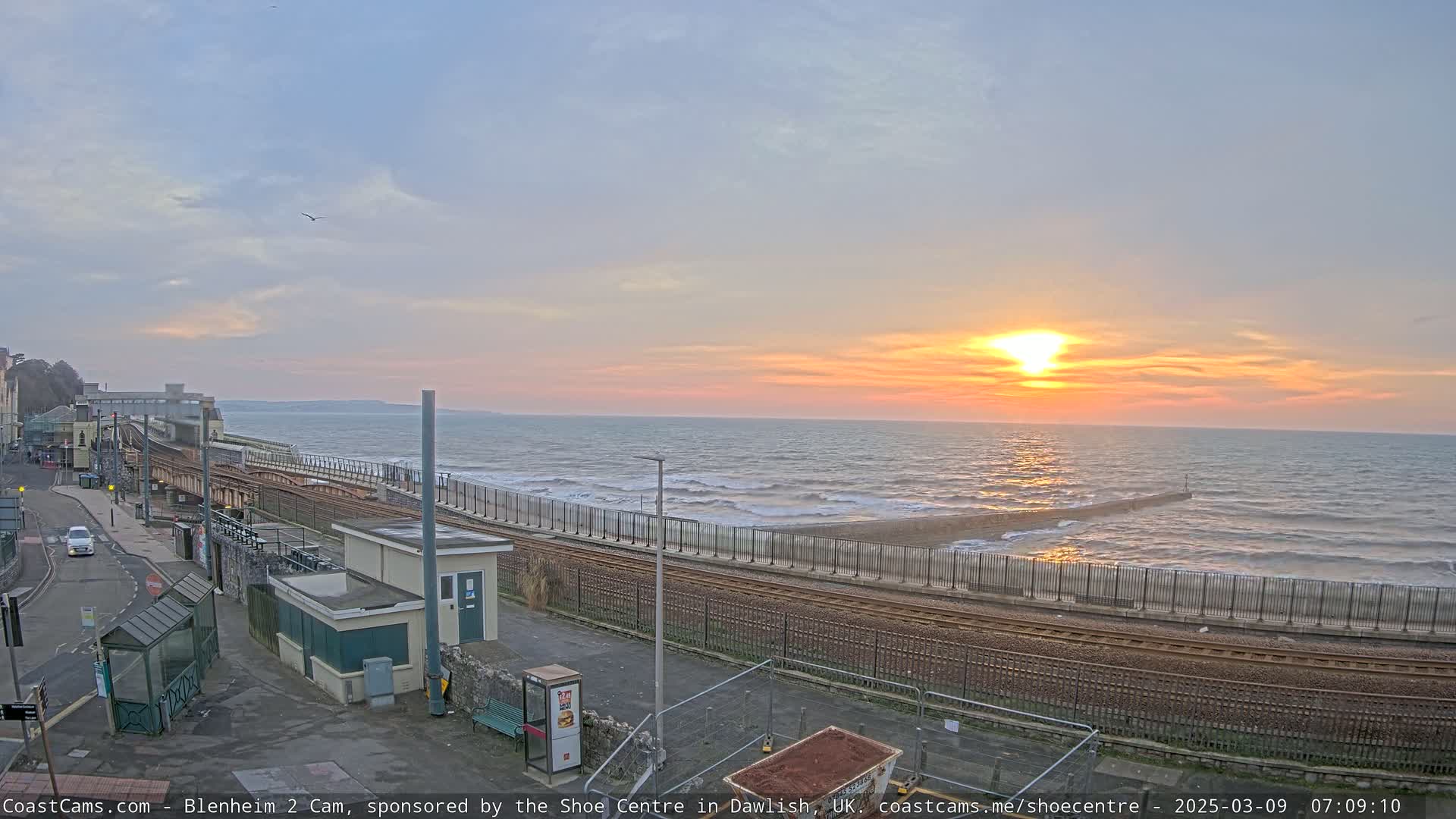 A coastal train track runs alongside a beach with a setting sun casting orange hues across a somewhat choppy sea.