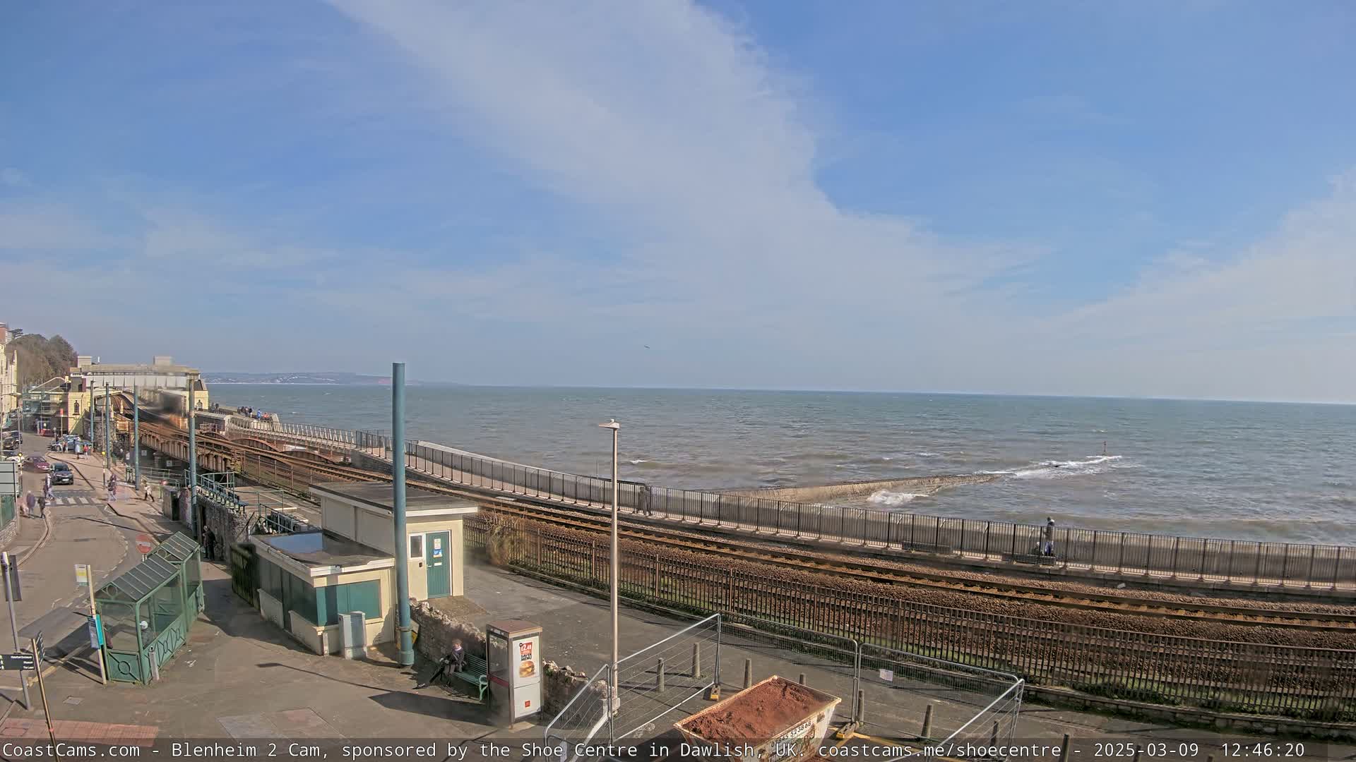 A seaside town scene on a partly cloudy day shows train tracks running alongside a seawall next to a calm ocean.