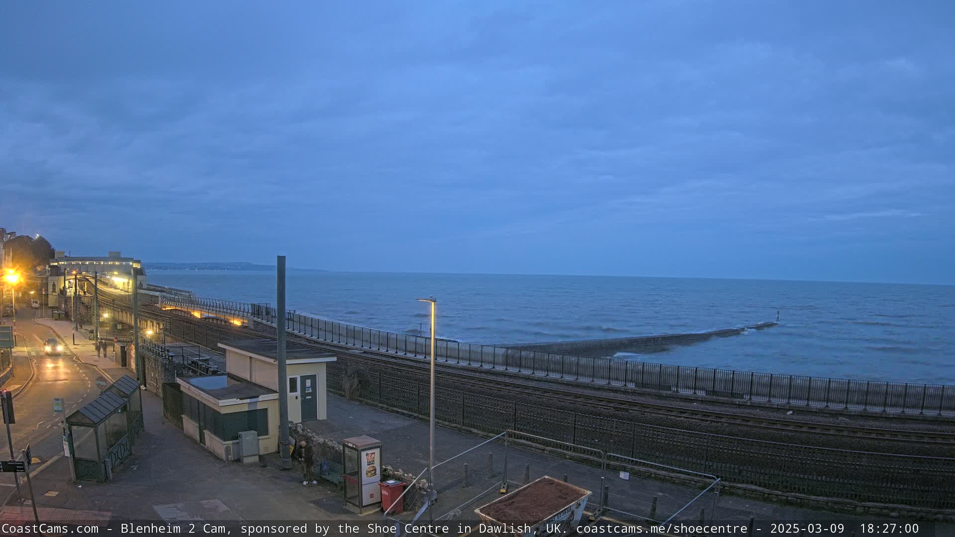 A coastal train track runs alongside a sea wall, with a calm, twilight sea visible in the background.
