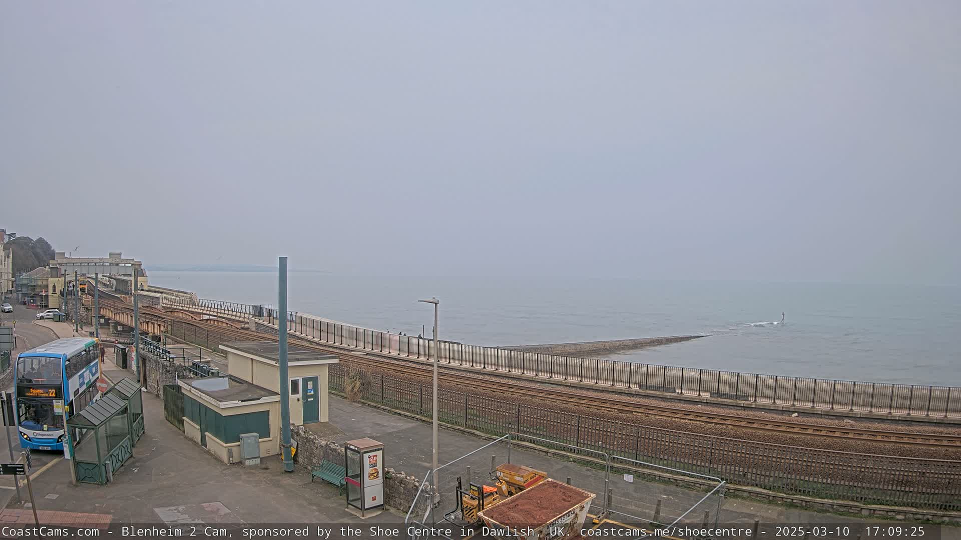 A hazy overcast day shows a coastal train track running alongside a sea wall with a bus and small buildings nearby.