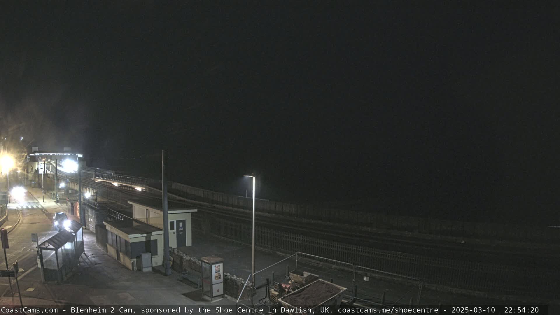 A nighttime view of a mostly deserted train station platform next to a dark body of water under a clear, dark sky.