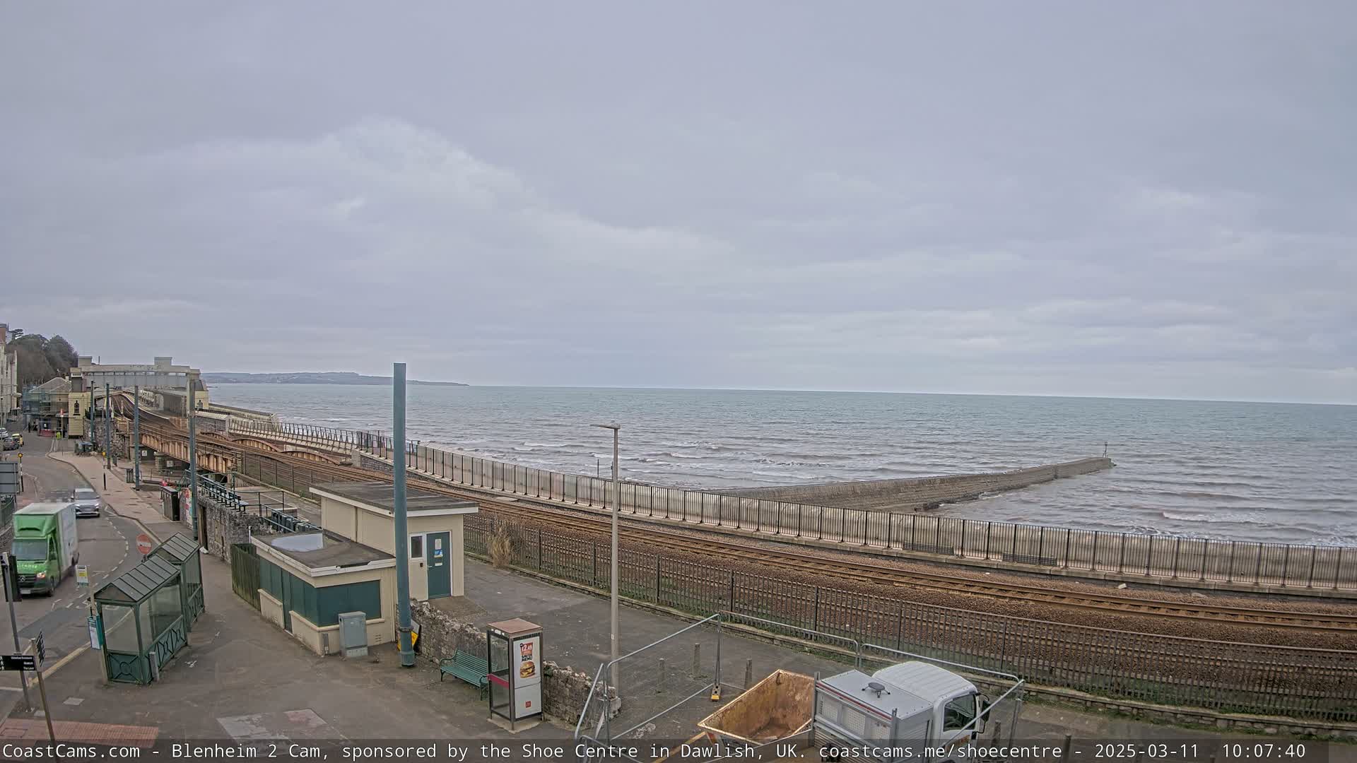 A coastal scene on an overcast day shows railway tracks running alongside a seawall that extends into a wavy ocean.