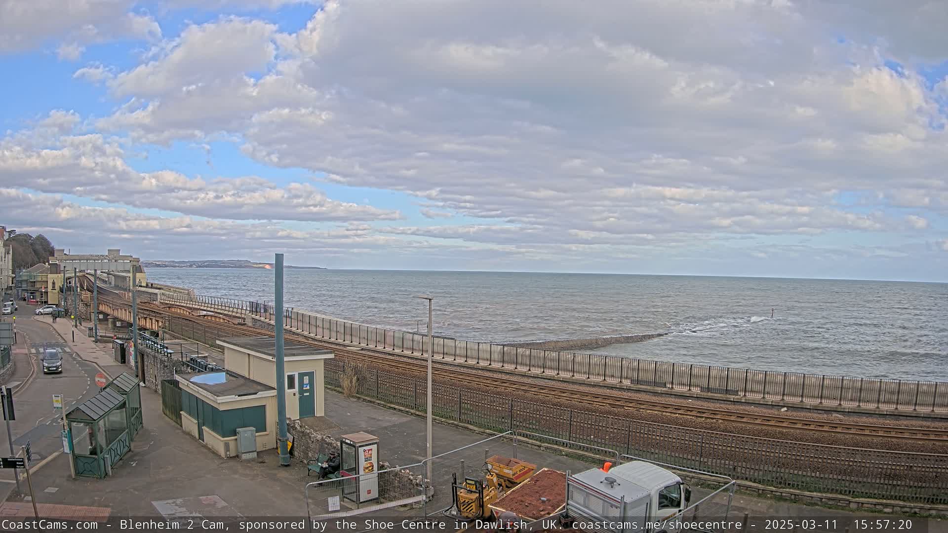 A partly cloudy day shows a coastal train track next to a sea wall with moderate waves crashing against it.
