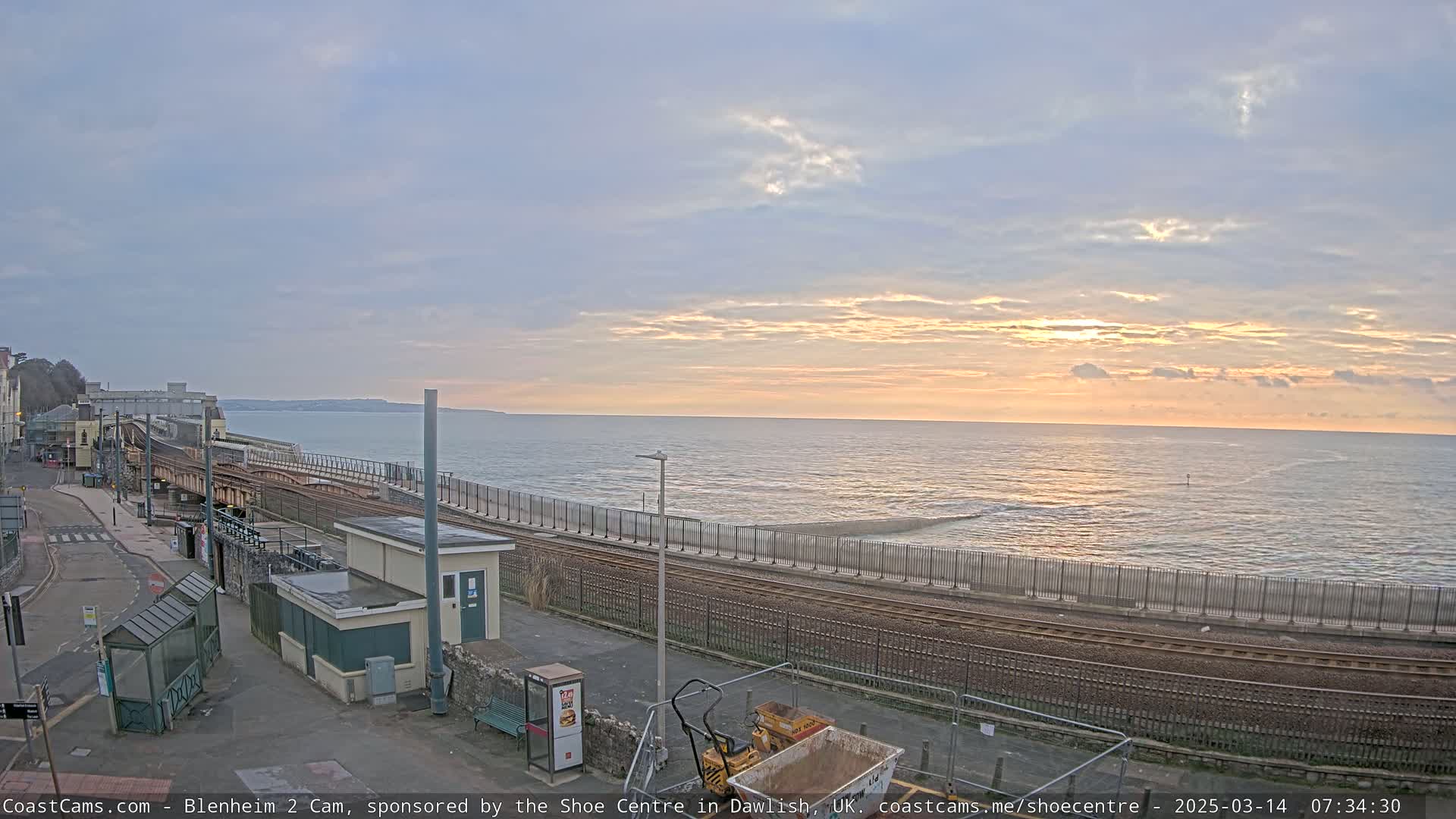 A sunrise over a calm sea, viewed from a coastal train station with a small construction vehicle present.