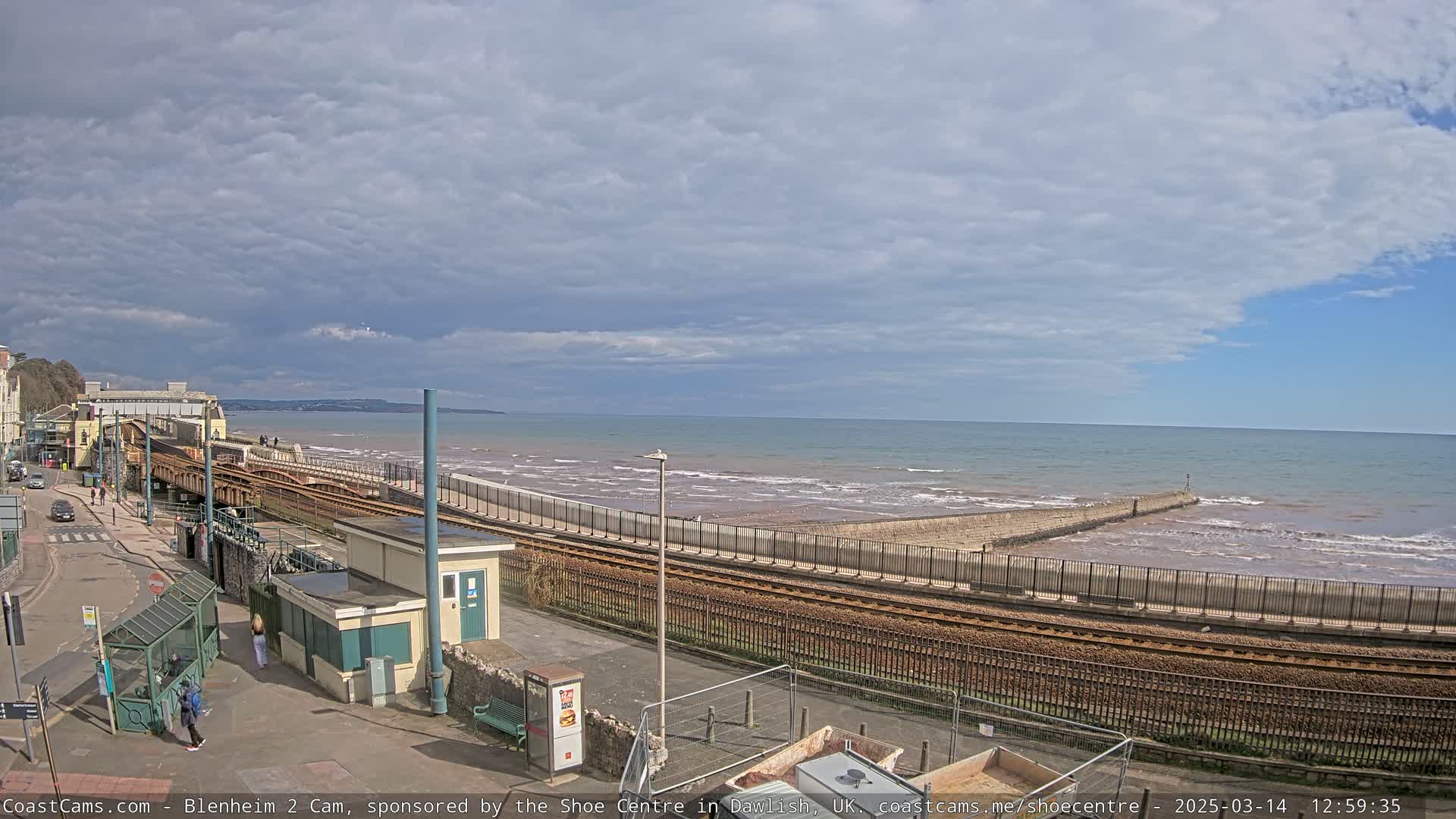A partly cloudy day reveals a coastal scene with a railway track running alongside a seawall and breaking waves.