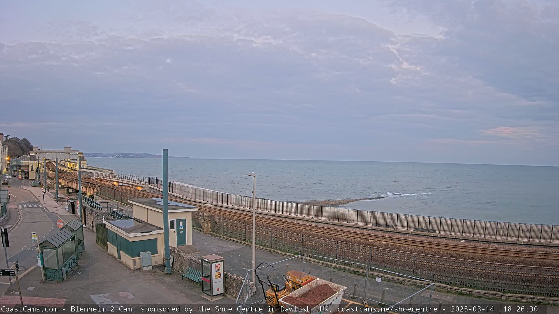 A train track runs alongside a calm sea under a cloudy sky, with a small building and construction equipment nearby.