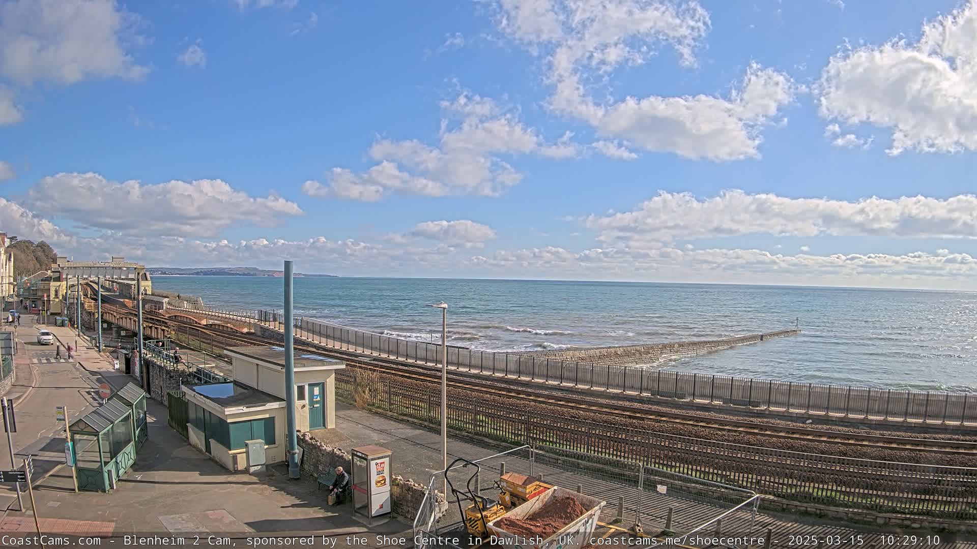 A partly sunny day shows a coastal train track running alongside a seawall with small waves breaking against it.