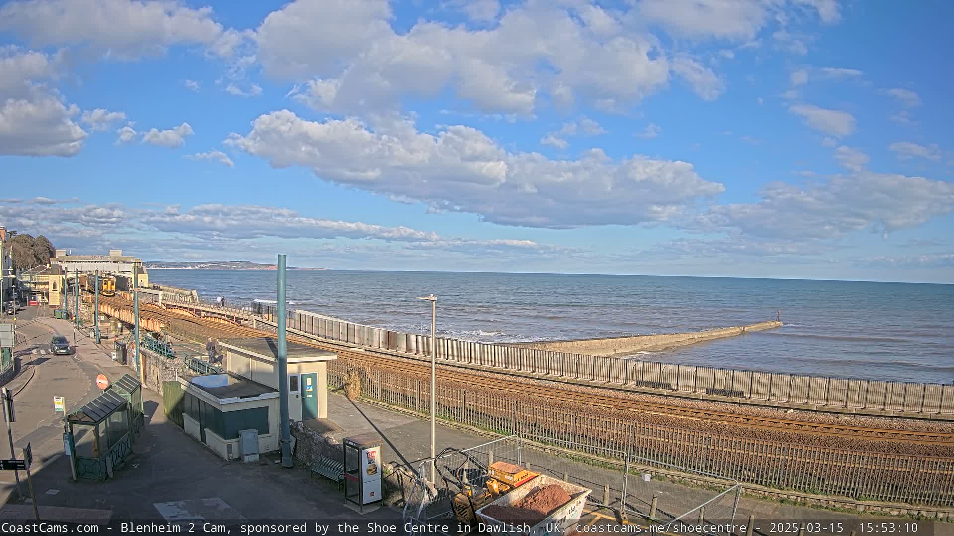 A partly cloudy day at a seaside train station with a yellow train visible on the tracks near a sea wall and calm ocean.