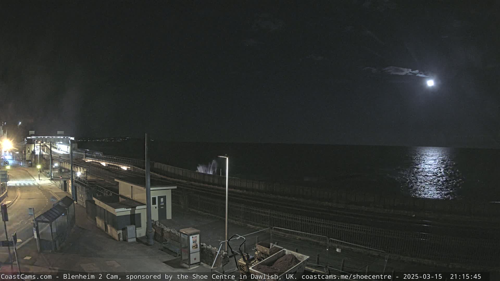 A nighttime view of a coastal train station and ocean under a bright moon.