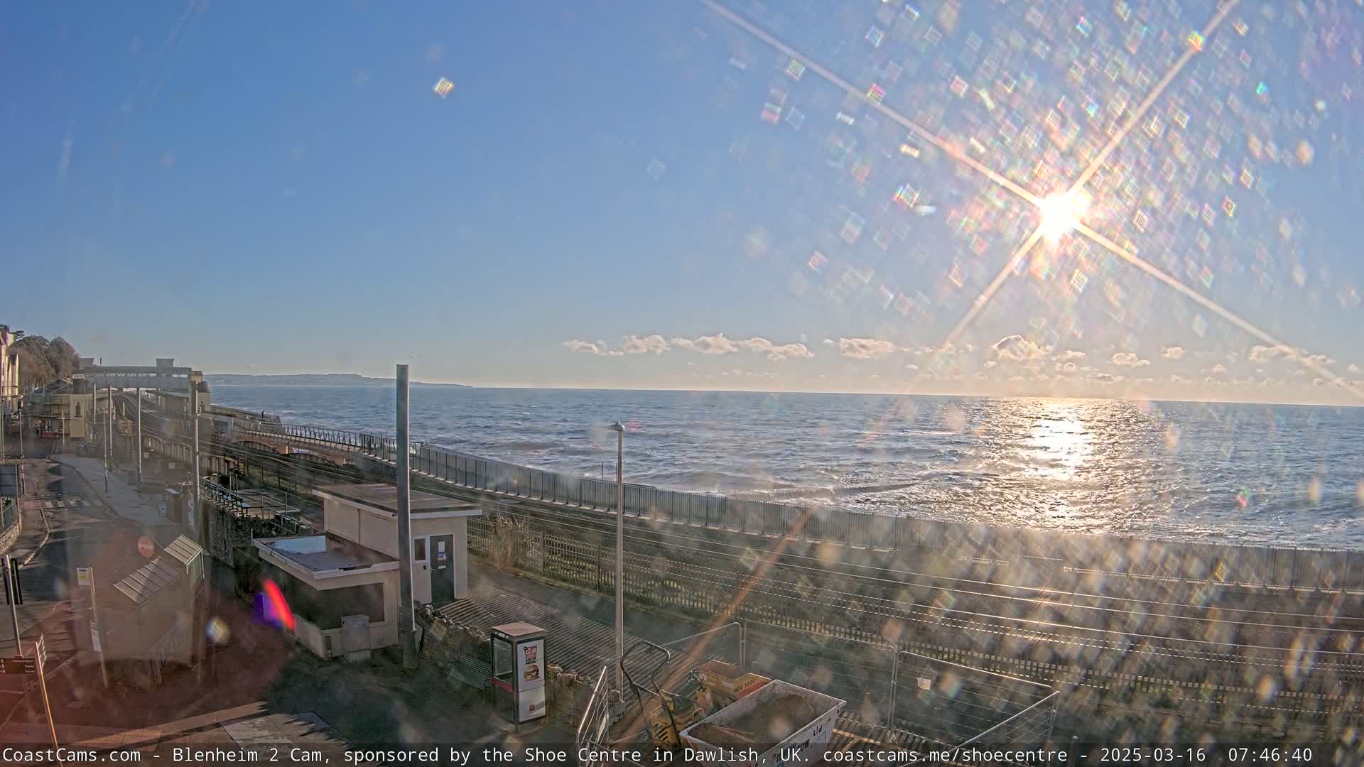 A sunny day shows a seaside train station overlooking a wavy ocean with the sun shining brightly.