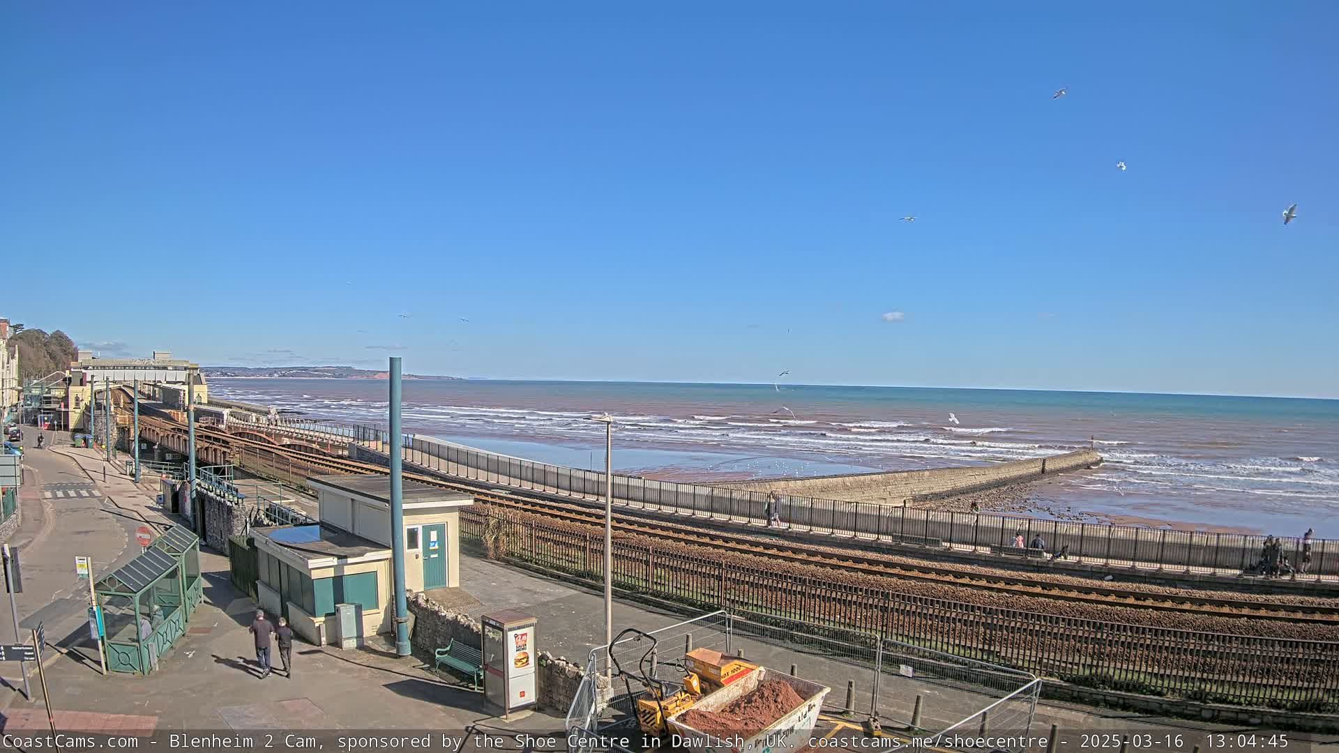 A seaside town on a sunny day shows a train track running along the shore, near a calm ocean with small waves, and construction equipment on the ground.