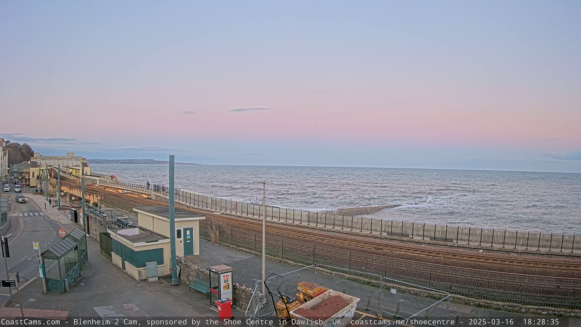 A seaside train track runs alongside a sea wall, with a calm sea under a pale pink and blue sunset sky.