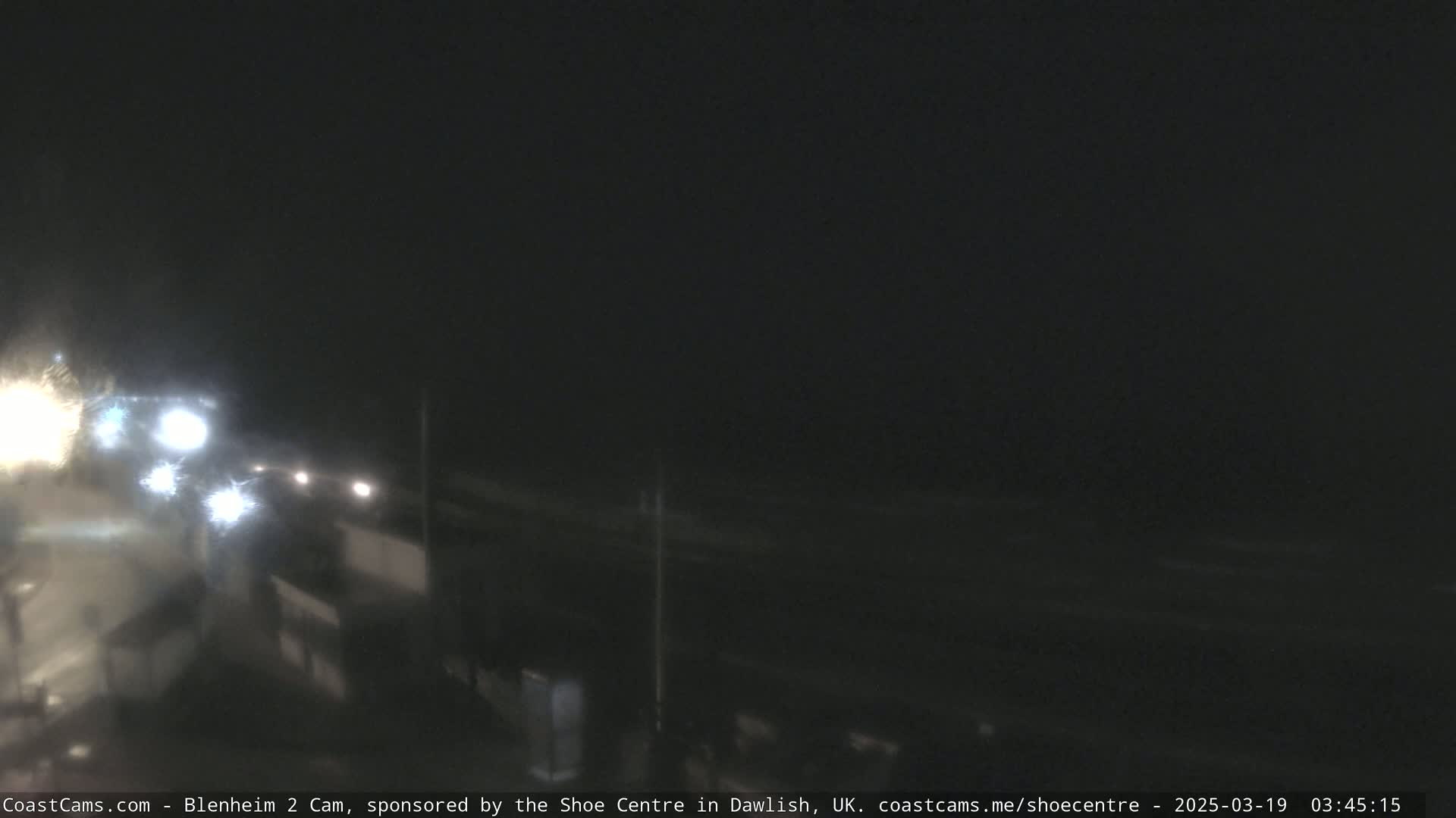 A nighttime, dimly lit coastal scene shows a dark ocean and a road with vehicle headlights visible in the distance.