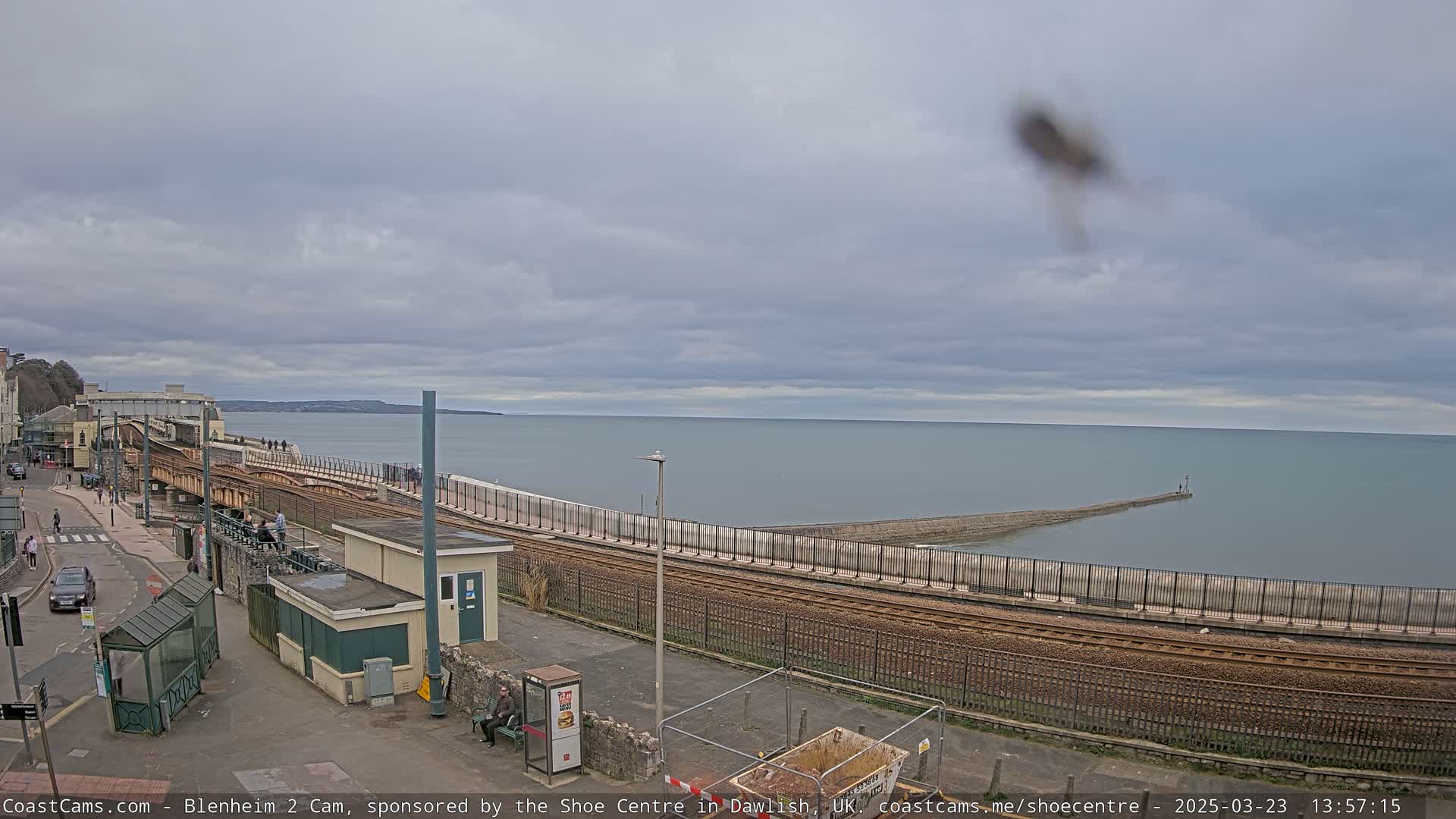 A coastal railway line runs alongside a sea wall, overlooking a calm sea under a cloudy sky.
