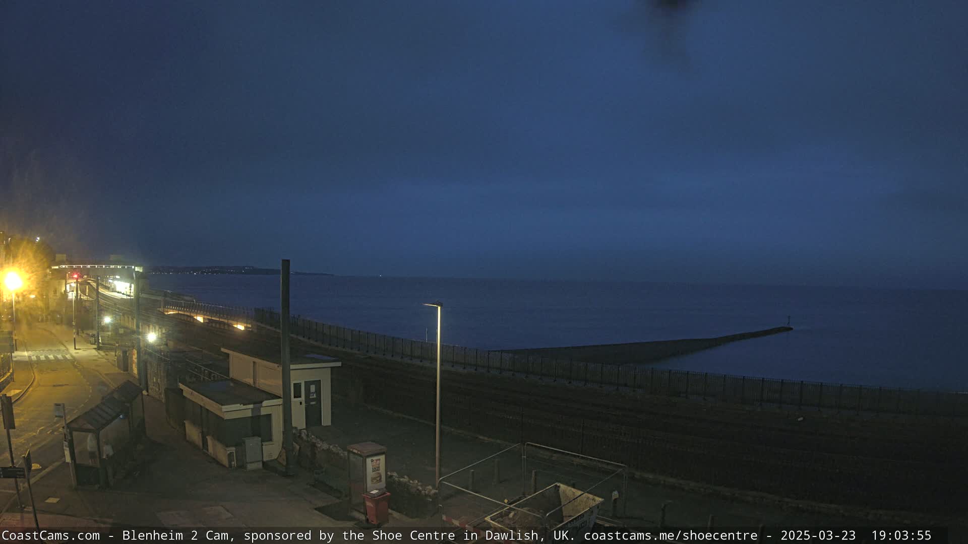A nighttime coastal scene shows a dark ocean, a breakwater, a seawall, and a street with minimal light.