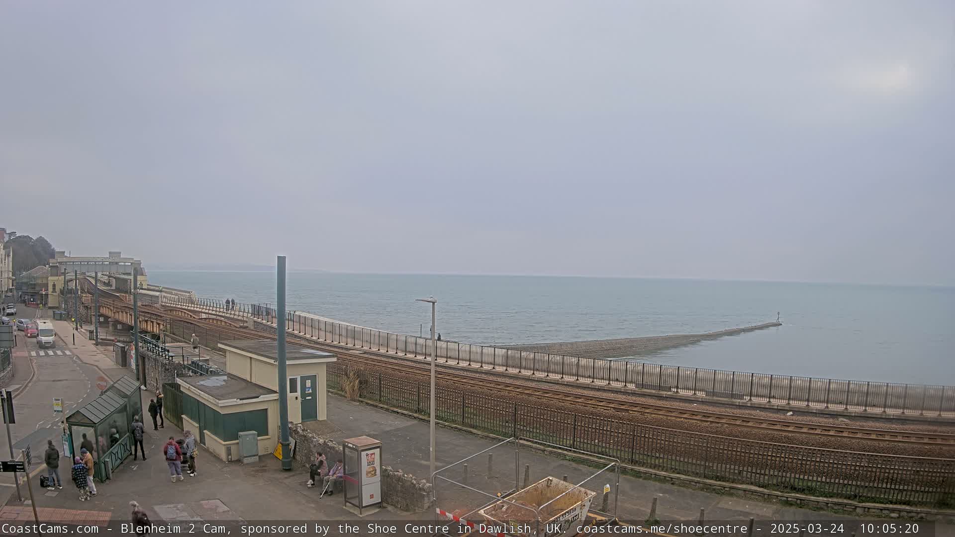 A coastal railway line runs alongside a sea wall, with people standing on a nearby paved area under an overcast sky.