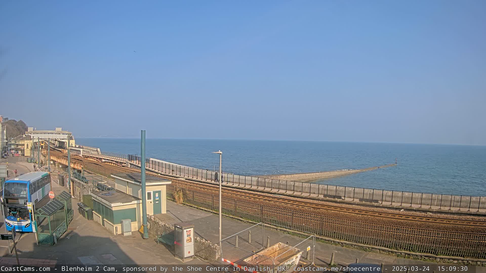 A clear, sunny day overlooks a coastal railway line next to a calm sea, with a bus parked nearby.