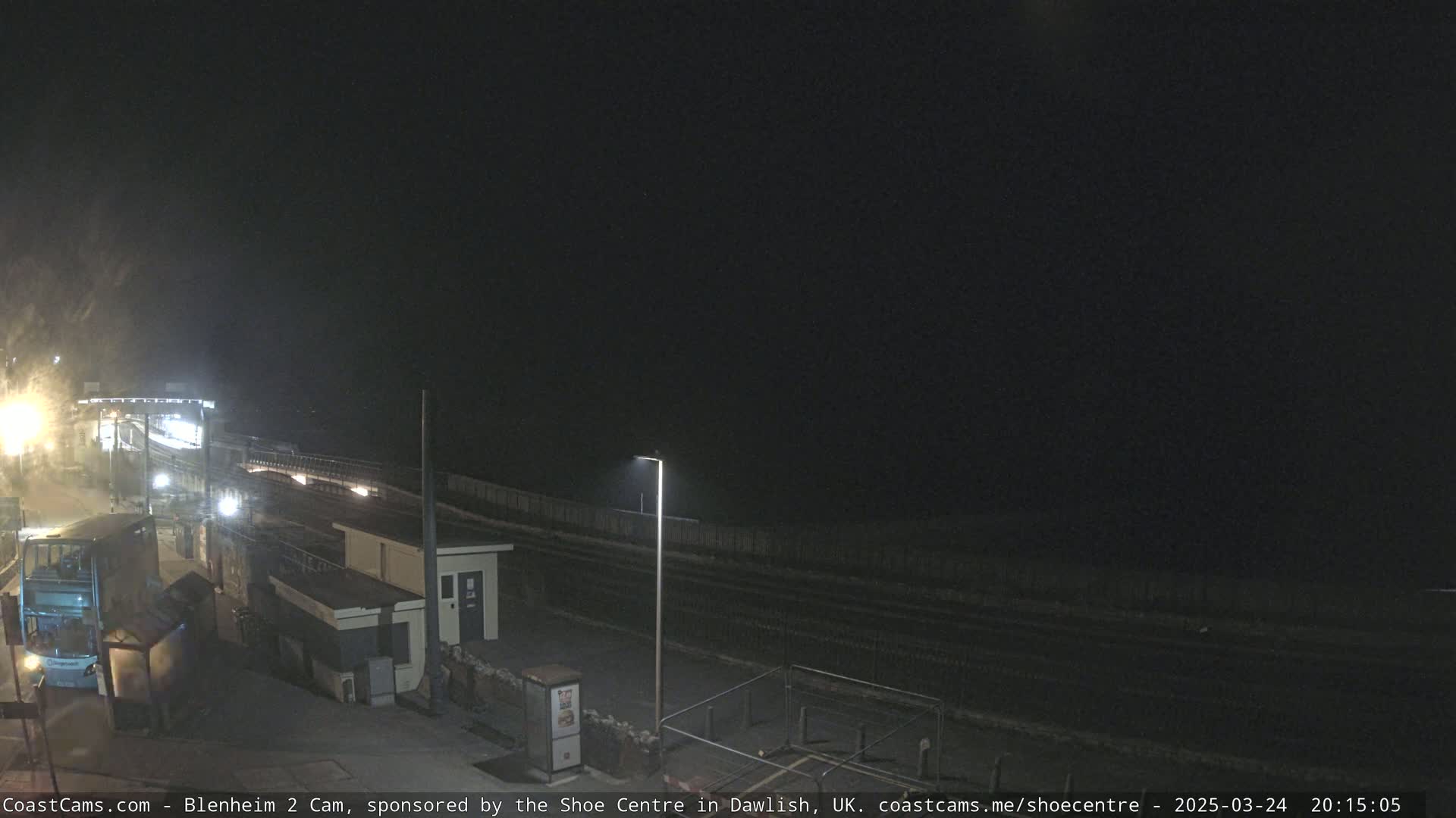 A nighttime view of a mostly empty train station platform next to a dark ocean under a clear, dark sky.