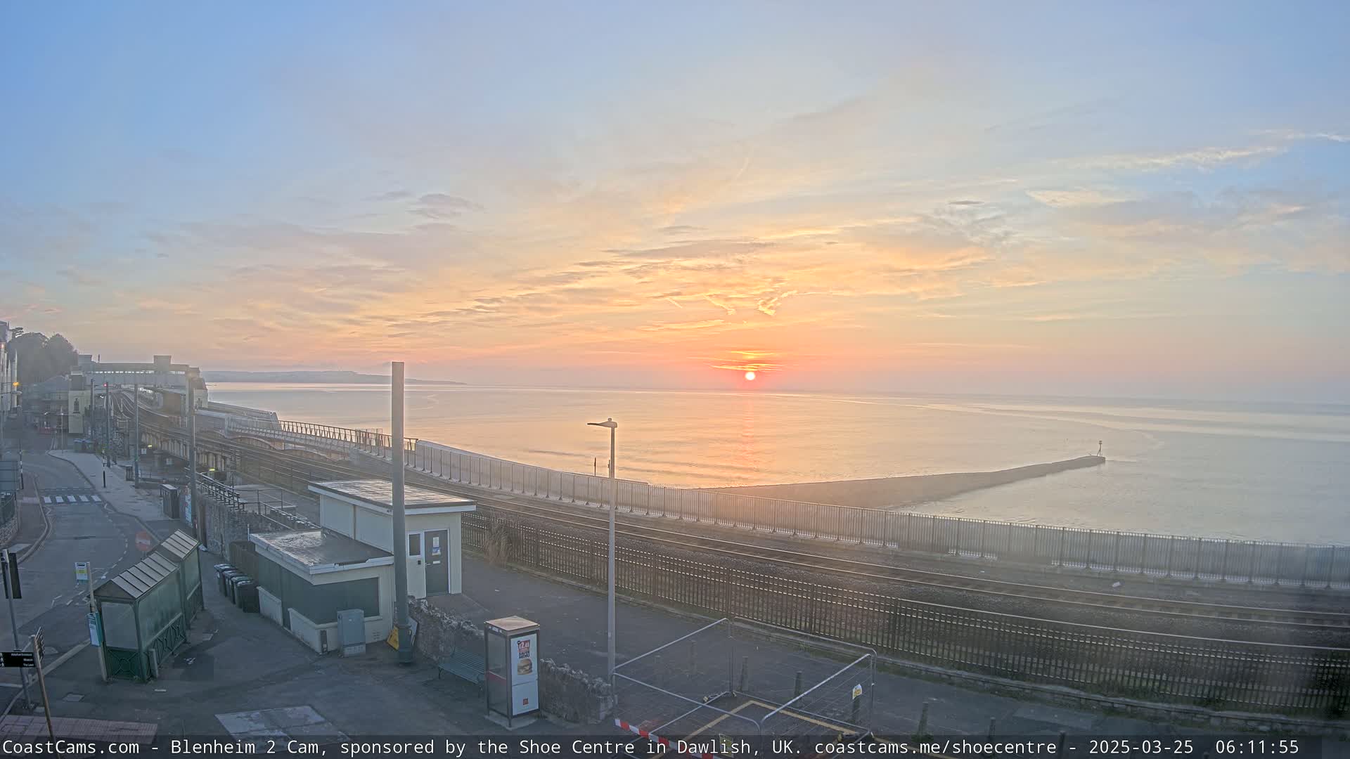 A seaside train station at sunrise, with a calm sea and a breakwater extending into the water.