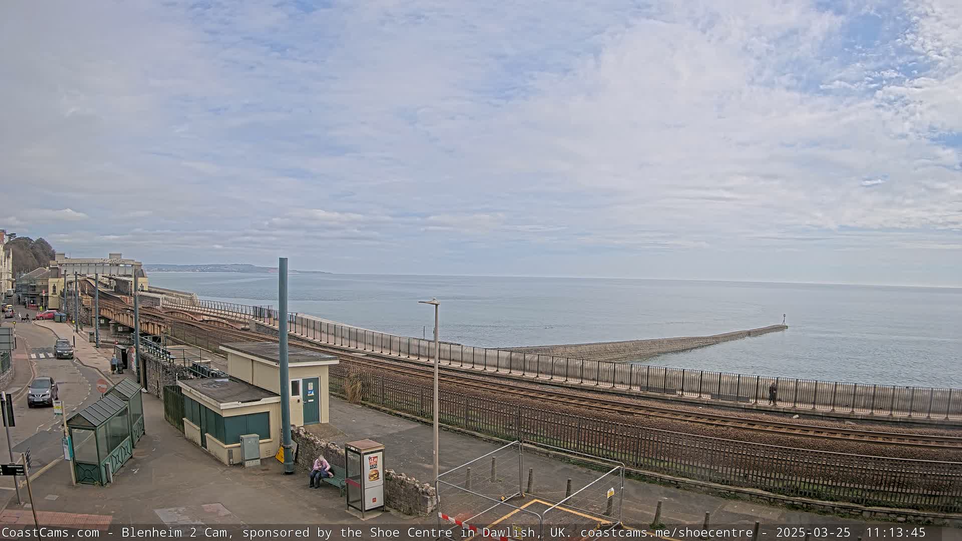 A coastal train track runs alongside a seawall separating it from a calm ocean under a mostly cloudy sky.