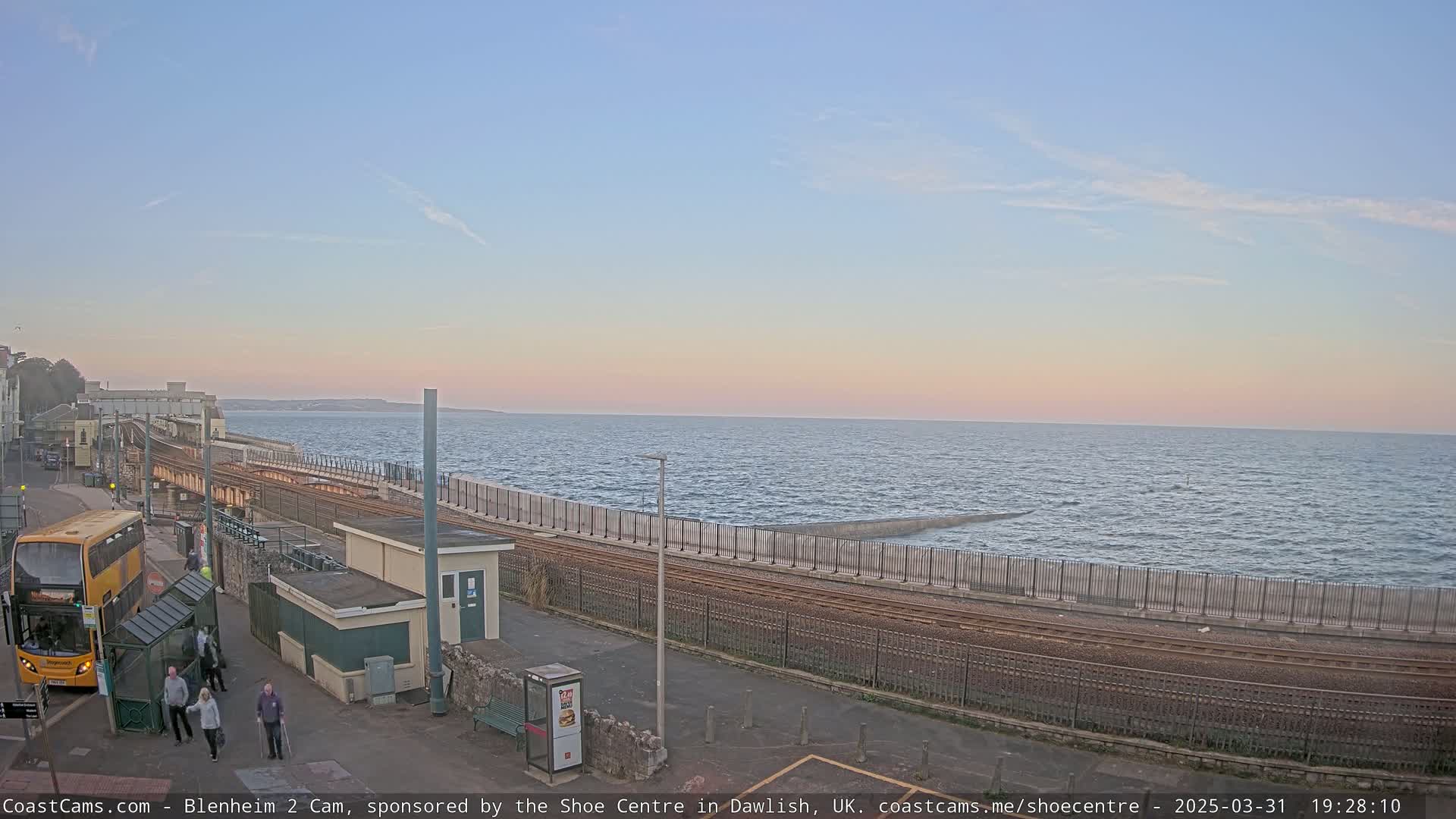 A seaside train track runs alongside a calm sea under a pale pink and blue sunset, with a bus and several people near a station.