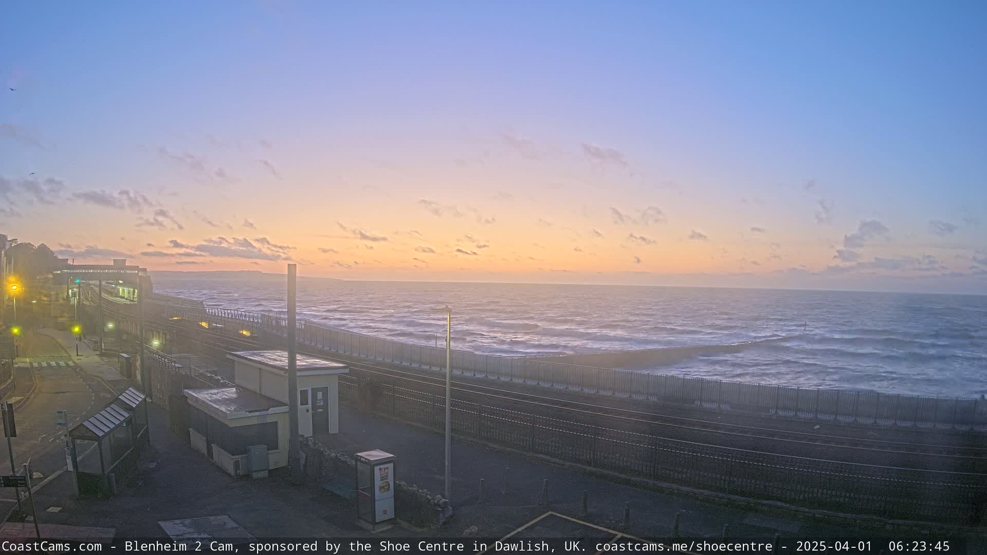 A coastal train station is visible at sunrise, with a choppy sea visible beyond a seawall.